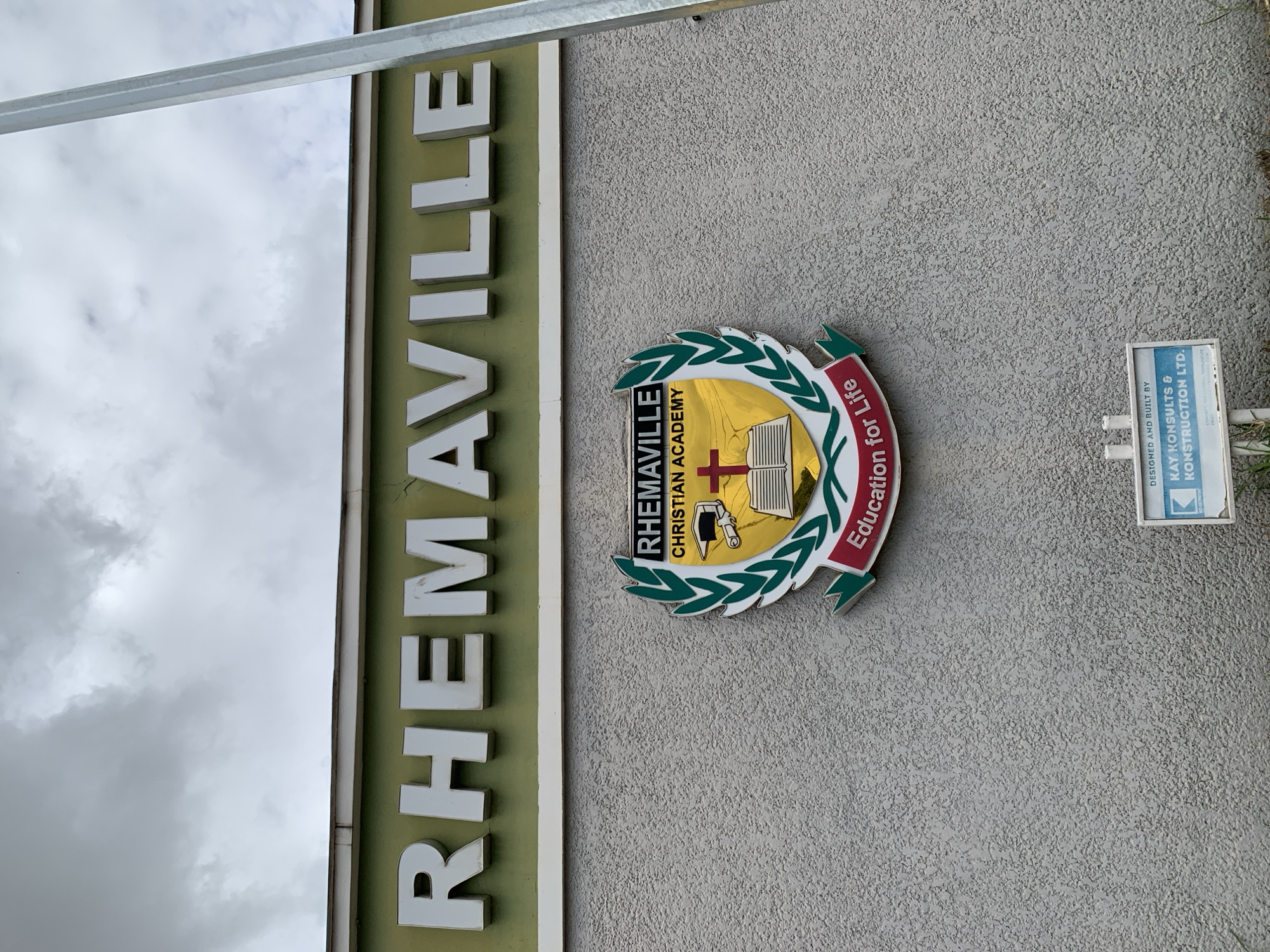 The image shows the exterior of a building with the name "RHEMAVILLE" displayed in large, white, raised lettering against a green background. Below the name is a crest with the words "RHEMAVILLE CHRISTIAN ACADEMY" and "Education for Life." The crest features a book, a graduation cap, a cross, and laurel leaves. The building's wall is textured and gray. In the lower right corner, a small sign reads "DESIGNED AND BUILT BY KAY KONSULTS & KONSTRUCTION LTD." The sky is overcast and cloudy. No people are visible in the image. This building appears to be Rhemaville Christian Academy, located in Jos, Nigeria.
