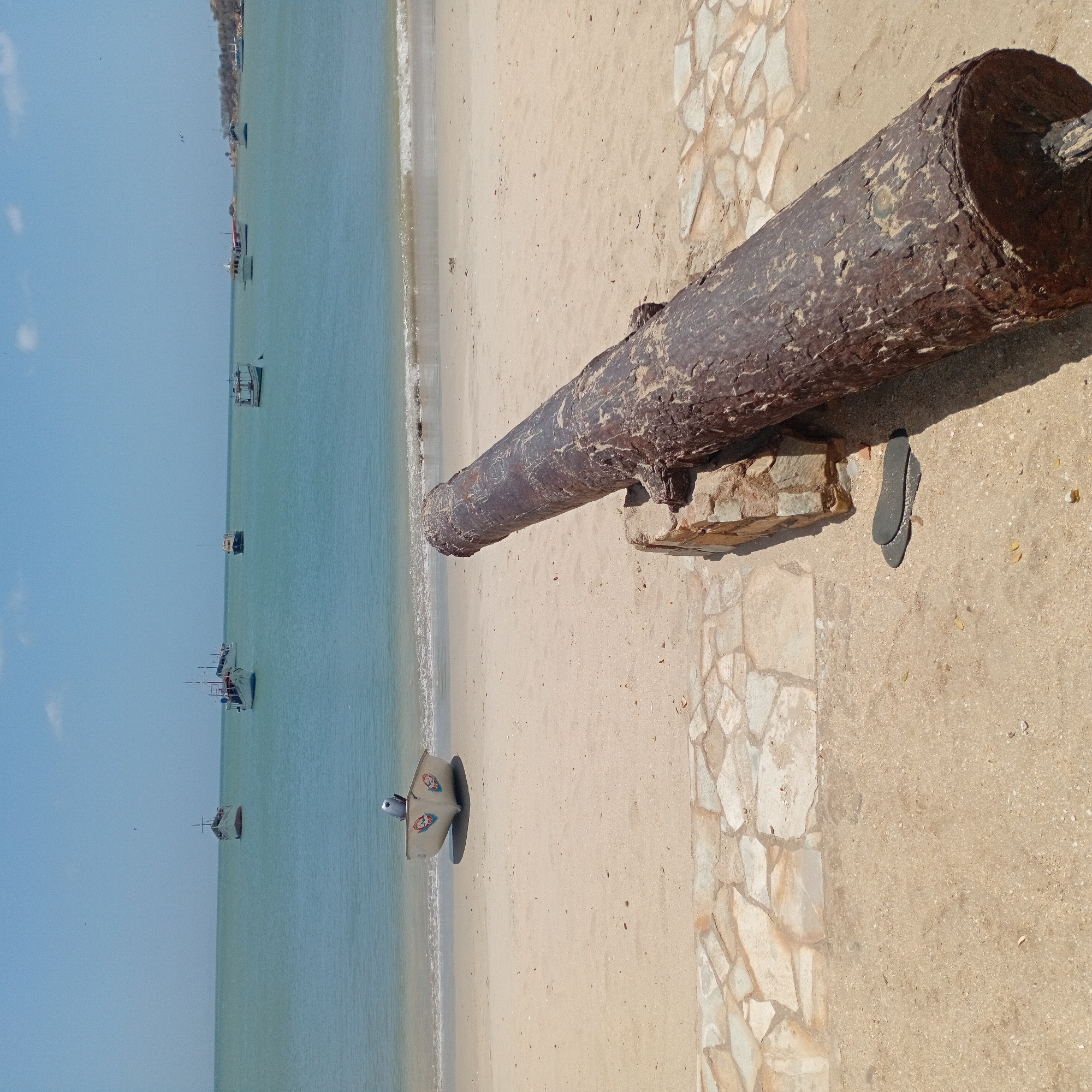 The image depicts a beach scene in an unknown city. A weathered, bronze-colored cannon rests on a stone support. Near the cannon, a single dark-colored flip-flop lies on the sandy ground. In the background, the turquoise ocean is visible, dotted with small boats. A small, white boat sits on the shore. A stone walkway borders the sand on the right. The sky is a clear blue.
