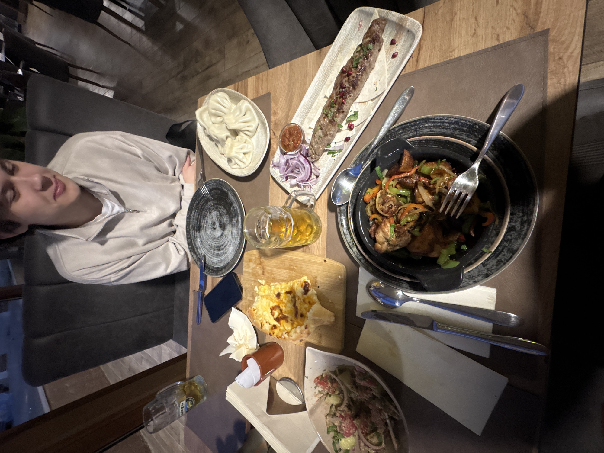 The image shows a person seated at a table laden with a variety of dishes. The table features a plate of dumplings, a long skewer of meat, a cast iron dish with vegetables and what appears to be roasted chicken, a dish with salad, and a piece of bread with cheese. There's also a beer glass, cutlery, a small condiment bottle, and a mobile phone on the table. The person is wearing a light-colored jacket and appears to be resting with eyes closed. The background suggests an indoor dining environment, possibly a restaurant. The location is not identifiable as Boreti, Montenegro without additional context.