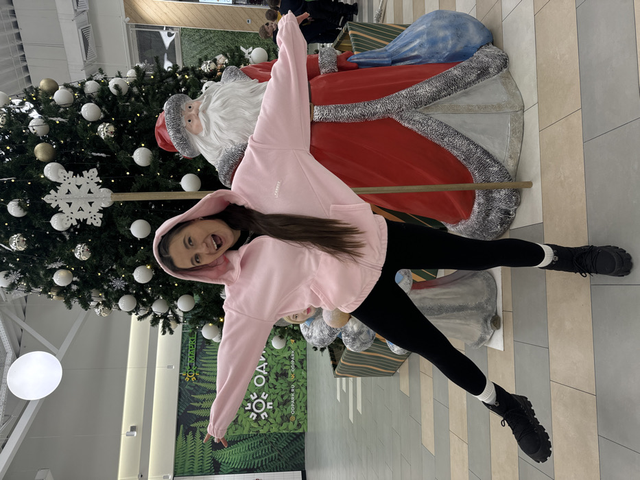 An indoor scene in a commercial establishment, likely a shopping mall or large store, in Yudino, Russia. A young adult female with long dark hair, wearing a pink hooded zip-up jacket with "FANZES" visible on the left chest, black leggings, and black boots with white socks, poses actively. She smiles widely at the camera with her mouth open, arms extended horizontally, and her left leg raised. She is positioned between a large decorated Christmas tree, adorned with white and gold spherical ornaments and a prominent white snowflake topper, and a large Santa Claus statue in a red suit with white fur trim, holding a blue sack. A smaller, lighter-colored statue resembling Snegurochka (Snow Maiden) is partially visible behind the female. These festive decorations are arranged within a low, striped planter box. The background features a large green wall graphic with fern patterns and a prominent circular logo displaying "ОДИН ПАРК" (ODIN PARK) and website addresses "ODPARK.RU" and "ODIPARK". The floor is tiled, and the high ceiling has visible structural elements and a large circular light fixture. Blurred figures of other individuals, including children, are present in the distant right background.