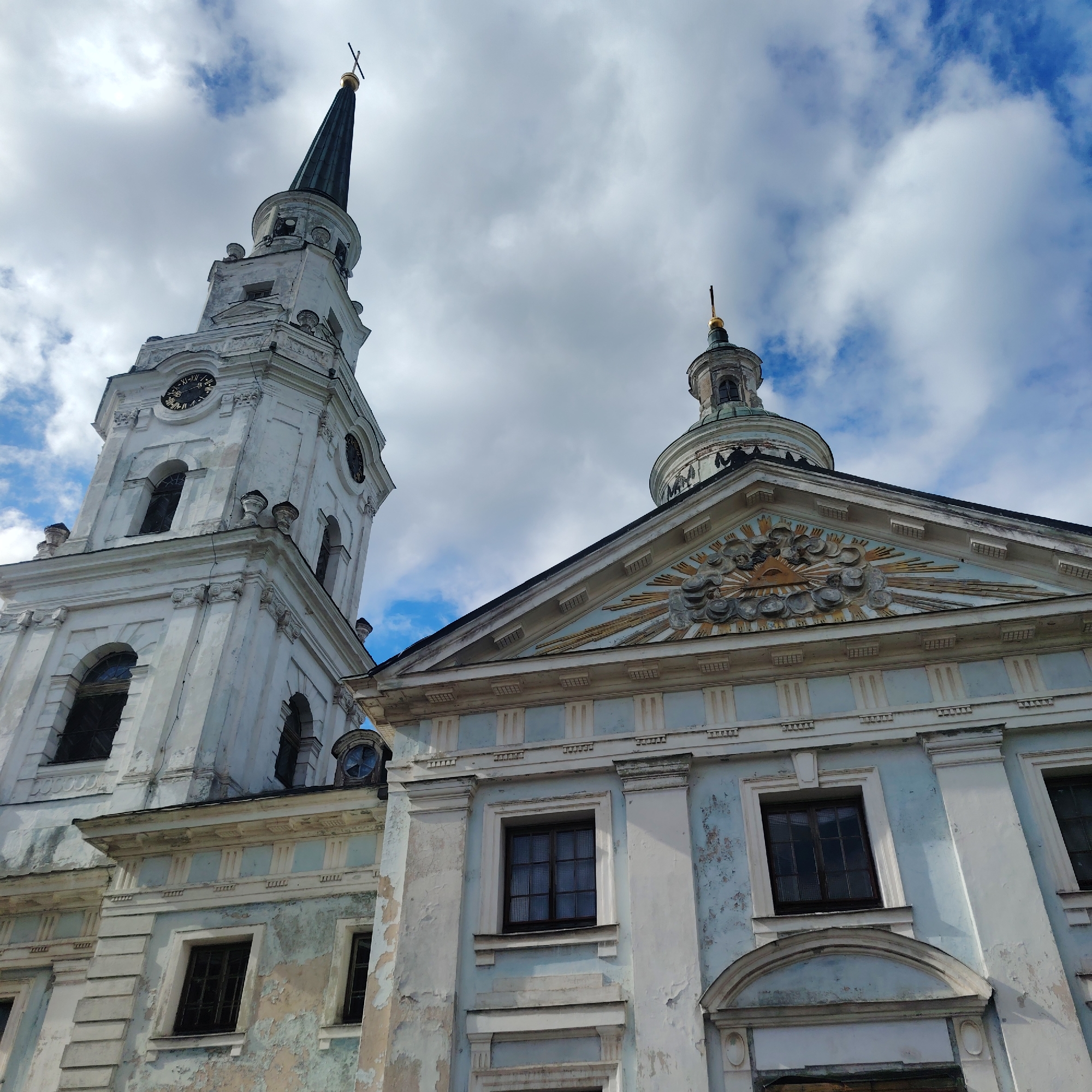The image depicts a low-angle view of two distinct architectural structures in Riga, Latvia, under a cloudy sky. The structure on the left is a tall, multi-tiered tower with a clock face and a spire topped with a cross. The structure to the right features a triangular pediment with ornate detailing, including a radiant sun emblem and a smaller domed tower with a cross on top. Both structures are pale in color with signs of aging and weathering, such as peeling paint and discoloration. Windows are visible on both structures. The overall impression is of historical buildings in a state of slight disrepair, juxtaposed against a dynamic sky.