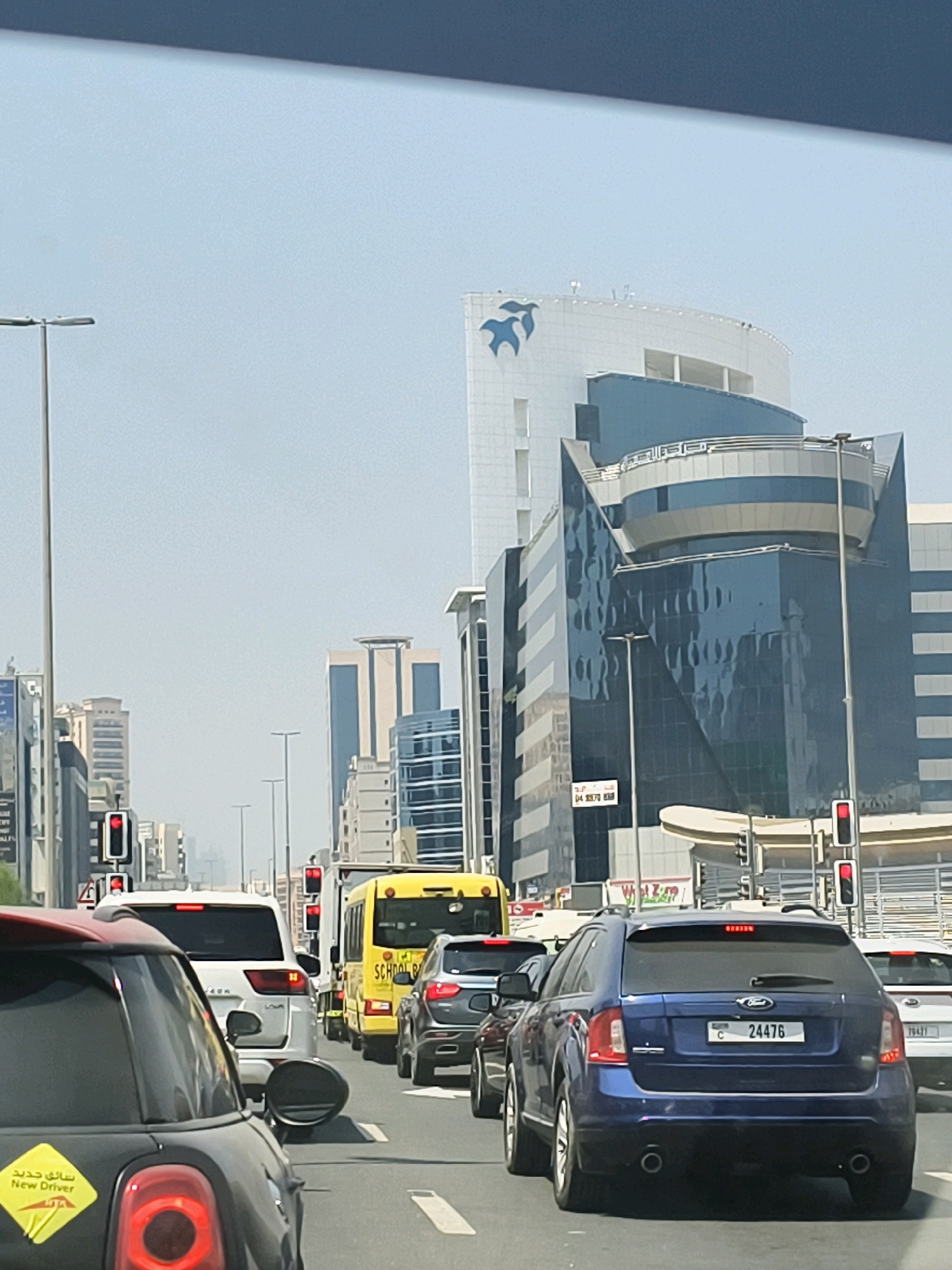 The image shows a busy city street in Dubai, United Arab Emirates, with multiple lanes of traffic. Several cars, including a blue Ford Edge with license plate C 24476, a white SUV, and a black Mini Cooper with a "New Driver" sticker, are visible. A yellow school bus is also in traffic. Tall, modern buildings with distinctive architecture line the street, including a large white building with a blue bird logo and a darker building with reflective glass. Traffic lights are visible, indicating that traffic is stopped. The sky is clear and bright.
