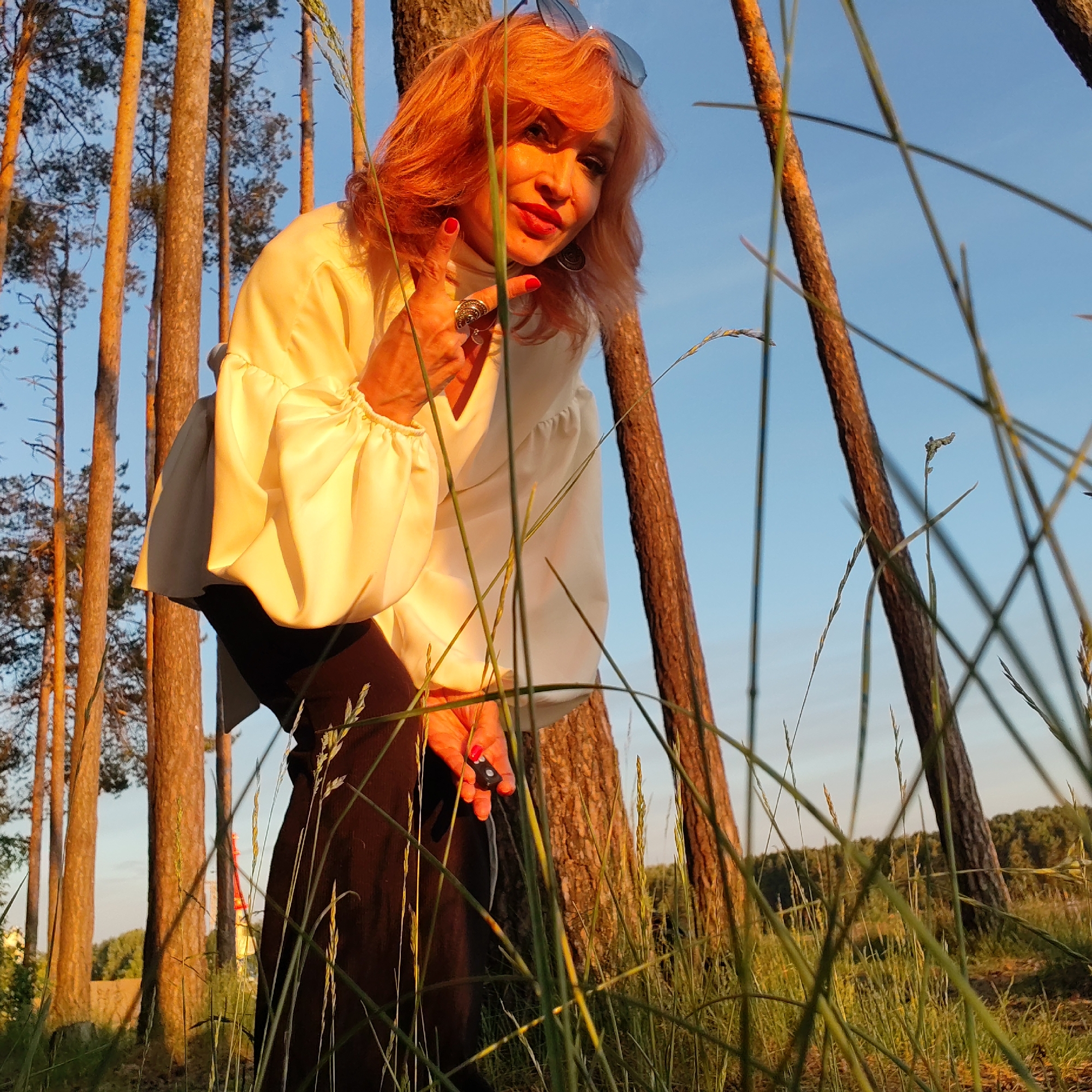 The image captures a woman posing in a sun-drenched pine forest, likely during the golden hour in Chaikovsky, Russia. The main subject, a woman with reddish-blonde hair and sunglasses resting on her head, is wearing a cream-colored, loose-fitting long-sleeved blouse and dark pants. She has bright red nail polish and a large ring on her left hand, which is raised in a gesture resembling a "rock and roll" or "shaka" sign. In her right hand, she holds a small, dark object.
She is leaning slightly forward, engaging directly with the camera with a confident expression, her red-painted lips prominent. The background features numerous tall, straight pine tree trunks, warmly lit by the setting sun, contrasting against a clear blue sky. The foreground is filled with green grass and various tall, dry stalks, some blurred, indicating a low-angle shot. The warm, soft light suggests it is late afternoon or early evening. The overall scene conveys a natural, outdoorsy setting with a stylish and spirited individual. No visible text is present in the image.
