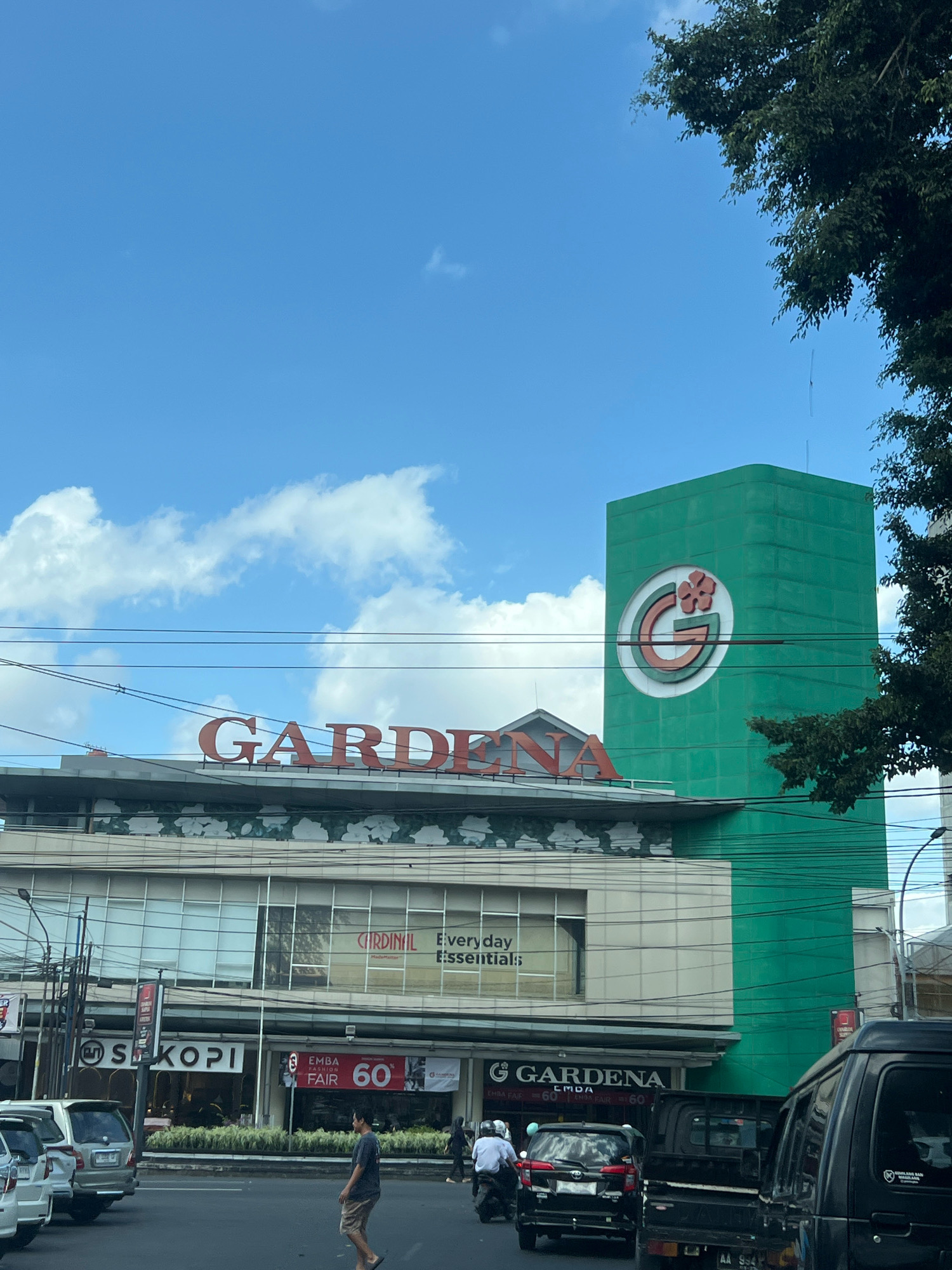 The image shows the Gardena department store in Magelang, Indonesia. The building is tall and green with the Gardena logo on the side. In front of the store is a street with cars and pedestrians. The sky is blue with some clouds. There is a tree in the foreground on the right side of the image. The Gardena department store is a well-known landmark in Magelang.