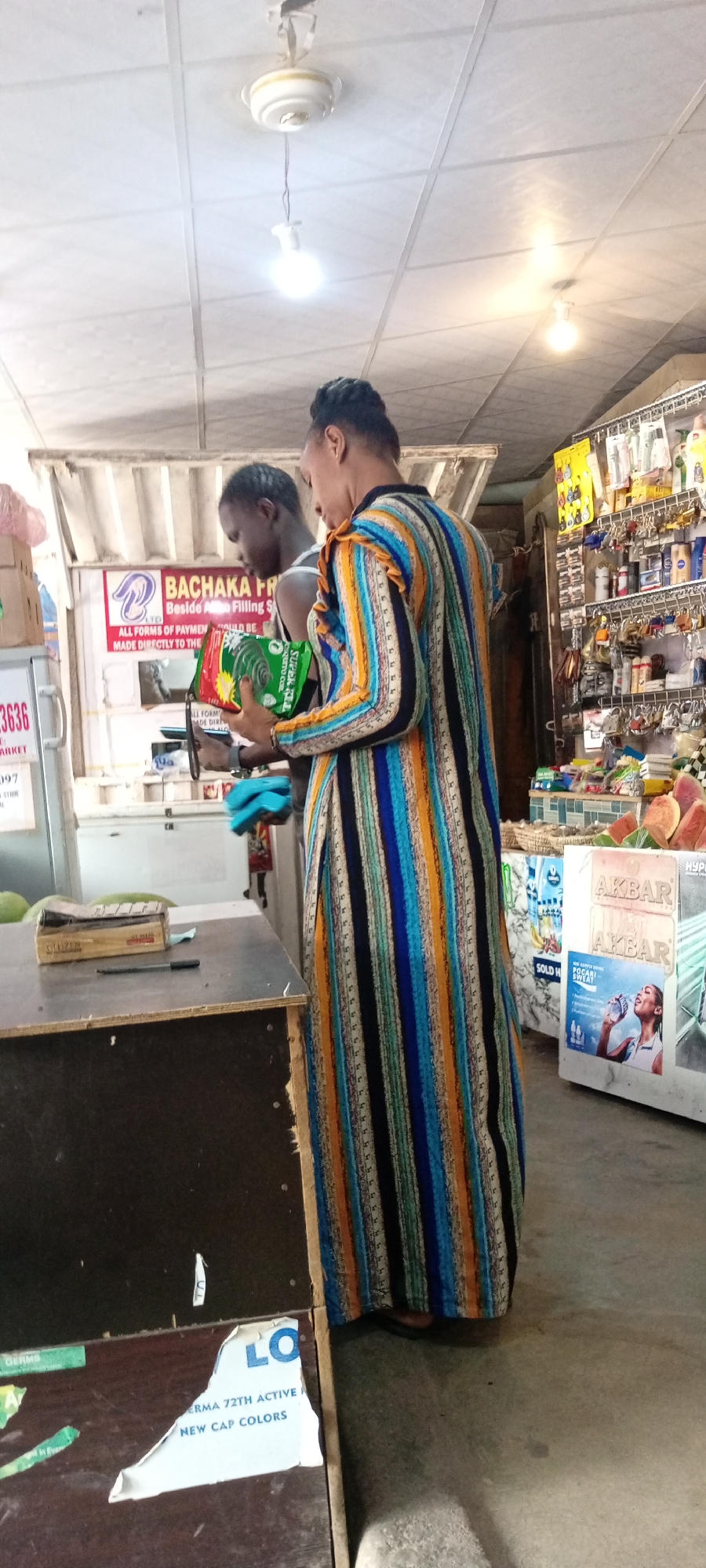 The image shows two people inside a store. One person, viewed from the back, is wearing a long, colorful, striped dress. This person is holding a green package of mosquito coils. The other person is partially visible and appears to be a younger individual. The setting is a small retail space, possibly a convenience store or market stall, with shelves stocked with various goods visible in the background. There's a counter in the foreground with some items on it, including a box and a pen. A ceiling fan and light fixture are visible overhead. Text visible in the image includes "BACHAKA FR", "ALL FORMS OF PAYMENT", "SOLD HA", "AKBAR", and "POCARI SWEAT". The overall impression is a busy, everyday scene in a retail environment in Nigeria. The time of day cannot be determined from the image.