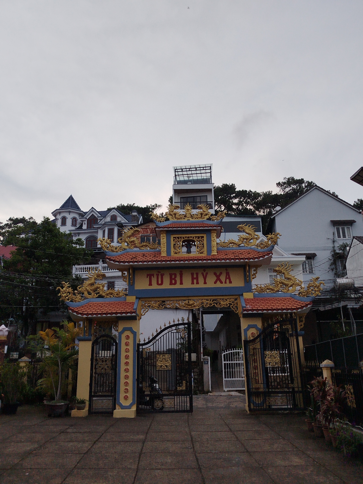 The image depicts a decorative gate in Da Lat, Vietnam. The gate is constructed with red tile roofing, yellow and blue painted columns, and ornate gold dragon accents. The words "TU BI HY XA" are prominently displayed above the gate opening. Behind the gate are several buildings of varying architectural styles, including a white multi-story house with a turret and a modern building with a glass-enclosed rooftop terrace. Utility wires are visible against a gray, overcast sky. The ground in front of the gate is paved with square concrete tiles and bordered by potted plants. A motorbike is parked near the gate. The scene conveys a blend of traditional and modern elements typical of the urban landscape.