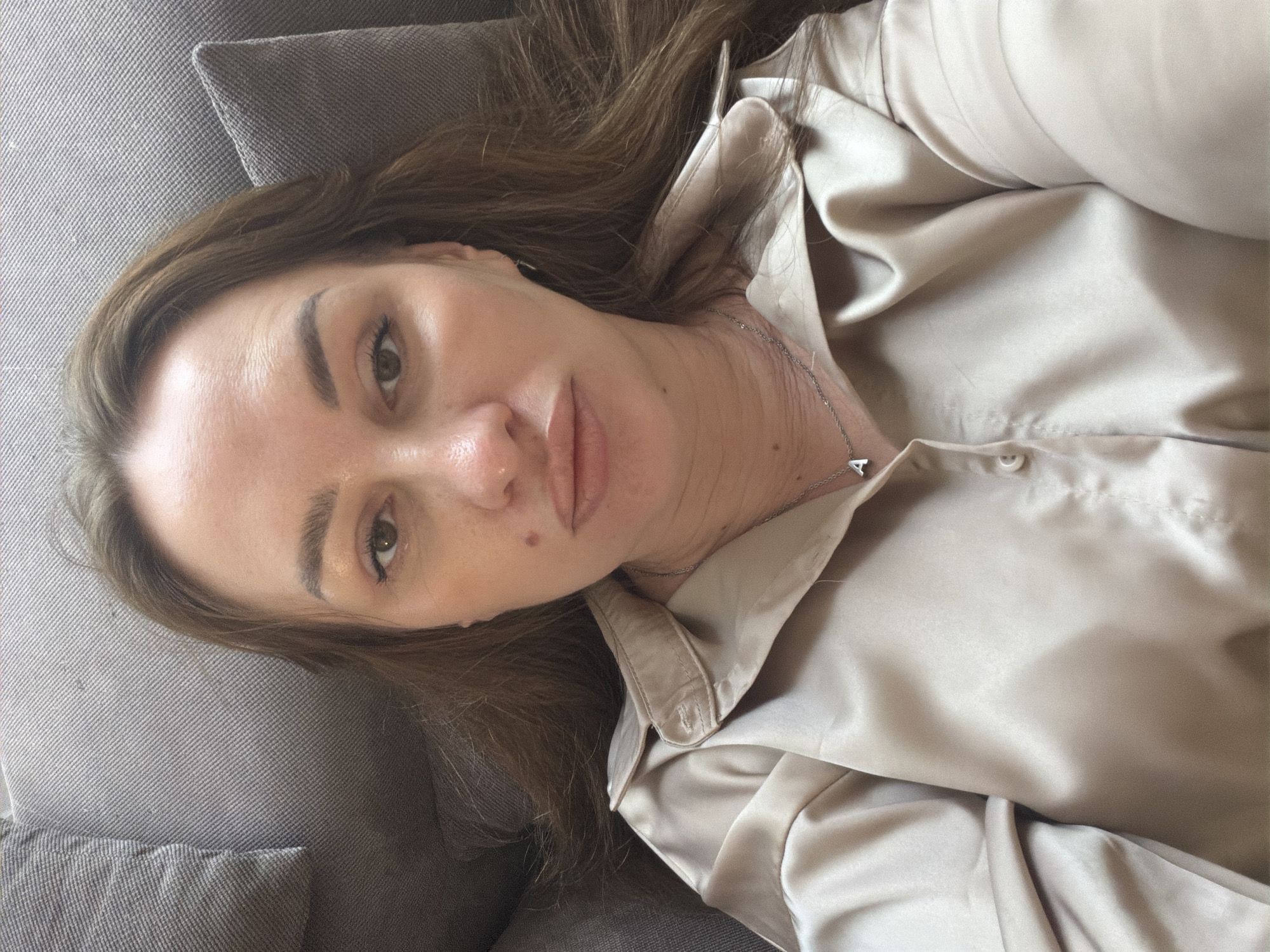 A medium shot selfie shows a woman lying on a grey couch in Malinovka, Russia. She has long, brown hair and is wearing a light-colored, silky button-down shirt. She is wearing a silver necklace with a small, silver "A" pendant. Her face is turned towards the camera, and she is looking directly at the lens. The lighting appears to be natural, coming from above. The background is the grey fabric of the couch and cushions. The image is taken from a slightly elevated angle.