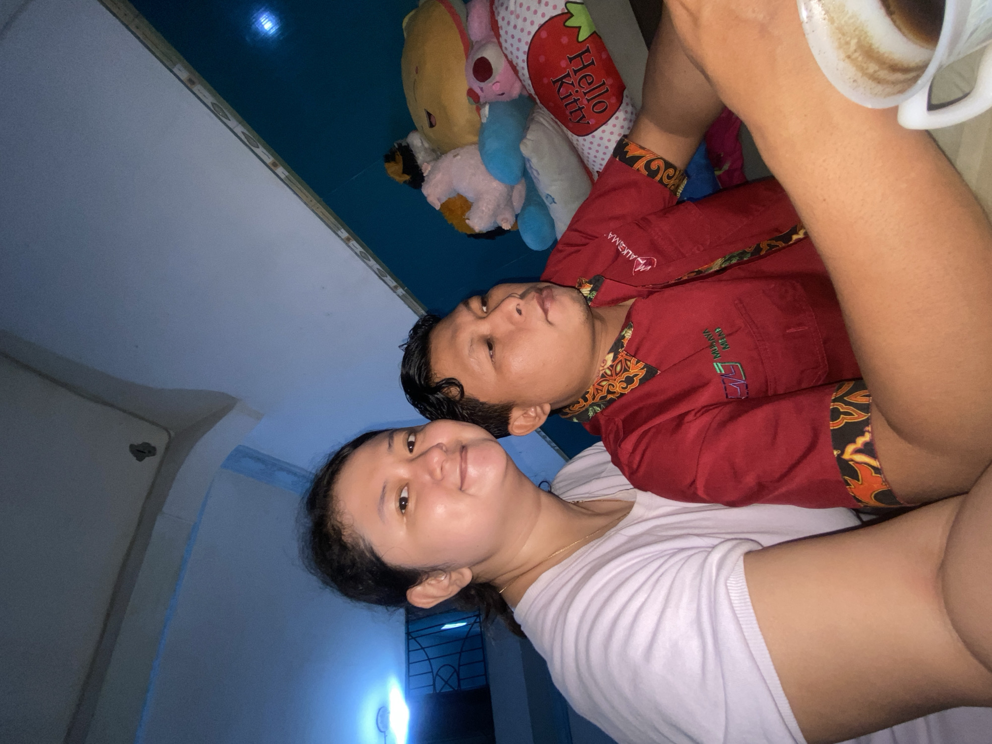 Two individuals, a woman and a man, are positioned in close proximity, likely sharing a moment. The woman, on the left, faces forward with a slight smile. She is wearing a light-colored t-shirt. The man, on the right, is looking to his left, away from the camera. He is dressed in a red polo shirt with patterned accents on the collar and sleeves, and a logo on the chest. Their arms are visible and appear to be resting on a surface. In the background, several plush toys are piled up, including a yellow bear, a pink and blue stuffed animal, and a pink bunny. To the right foreground, a white cup filled with a dark beverage, presumably coffee, is partially visible. The lighting in the scene is dim, with a blueish tint emanating from an unseen light source, possibly from a television or other electronic device, suggesting an indoor setting, potentially a living room or a communal area. The environment suggests a comfortable, possibly domestic, atmosphere. The specific location in the city of Gelam, Indonesia, cannot be determined from the visual information provided.