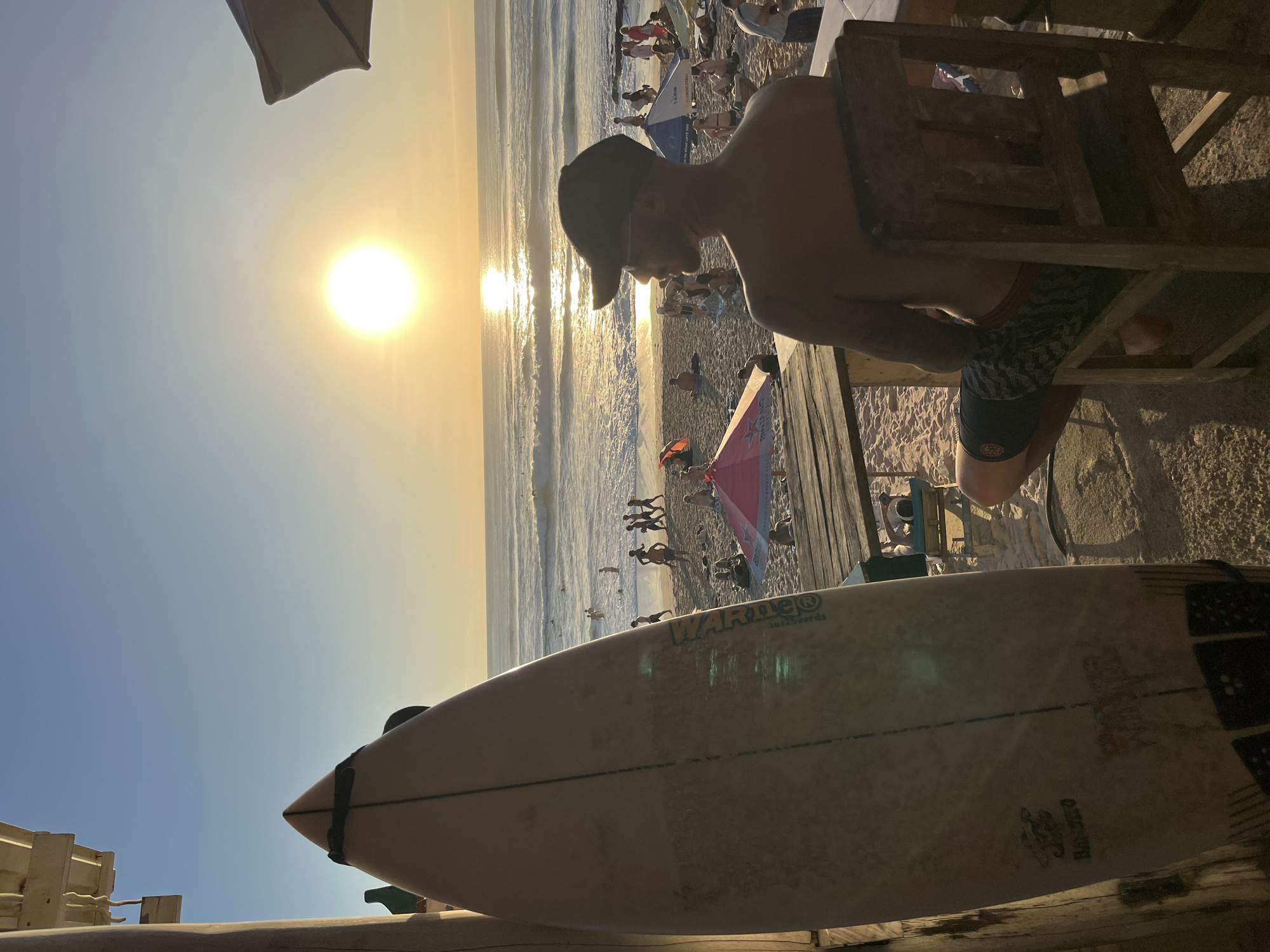 A man wearing a black baseball cap and patterned swim trunks sits in a wooden chair on a beachside wooden platform, facing the ocean. A surfboard is propped up on the left side of the frame in the foreground. In the background, people are on the sandy beach and in the water. The sun is setting over the ocean, casting a warm glow on the scene. The location is Kuta Selatan, Indonesia.