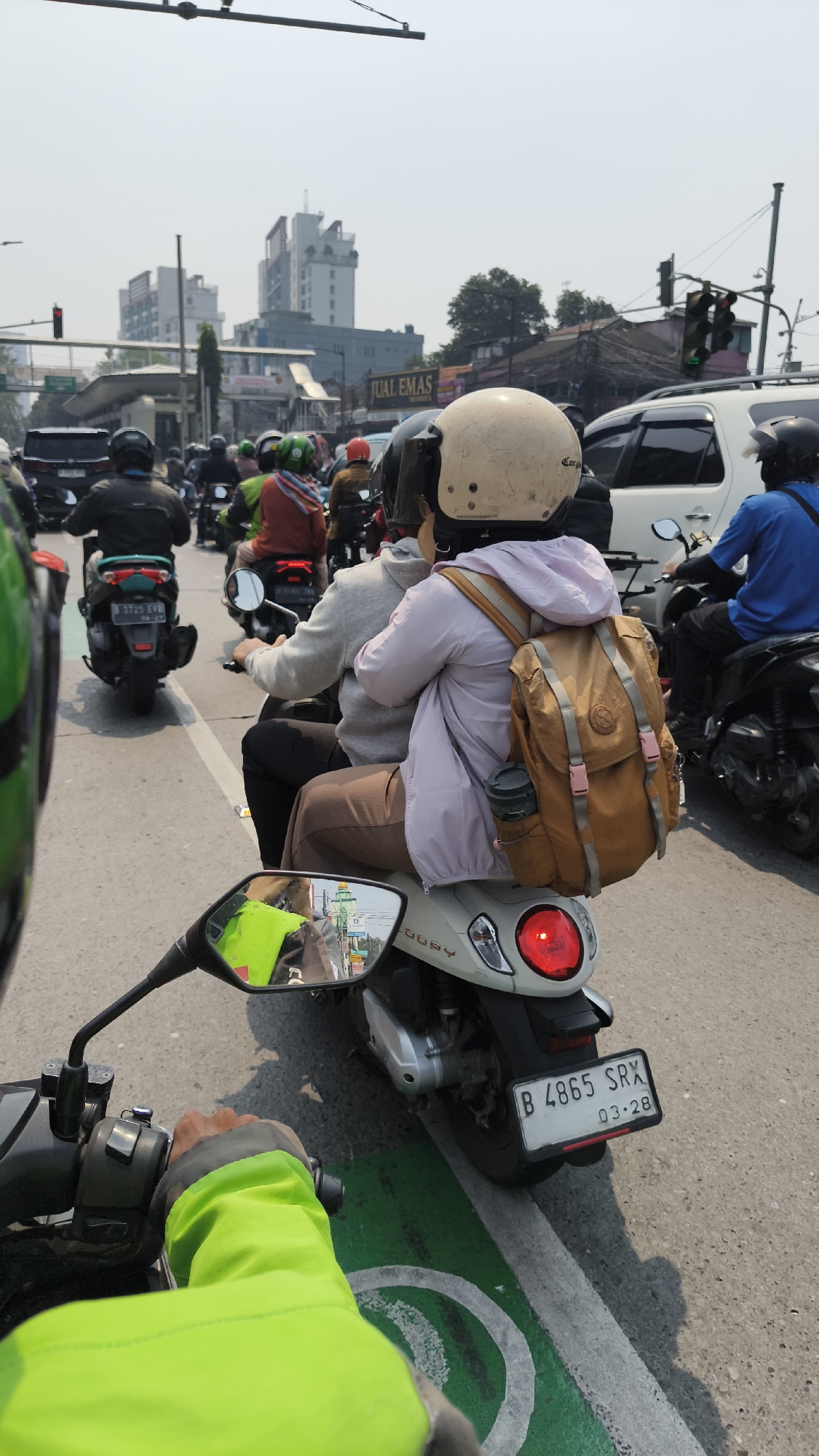 The image captures a congested street scene in South Jakarta, Indonesia, likely during daylight hours based on the bright sunlight and clear sky. The foreground shows the perspective of a motorcycle rider, with their lime green jacket and hand visible. To the right, another motorcycle's rear is visible, featuring a white scooter with a red taillight and a license plate reading "B 4865 SRX 03-28".
In the middle ground, several motorcycles and their riders are navigating the traffic. A prominent subject is a motorcycle with two passengers. The passenger in the back, wearing a light purple hoodie and a tan backpack, is the most visible. They are seated behind the driver, who is wearing a light-colored helmet and a grey hoodie. The driver's head and shoulders are visible. The scooter they are on is white and has "LOBBY" written on its side.
Further back, other motorcycles are interspersed with cars. A white car is seen to the right of the main subjects, and a black SUV is to the left. The traffic appears to be moving slowly, suggesting a traffic jam.
The background reveals the urban environment of South Jakarta. Buildings of varying heights are visible, including multi-story structures with balconies and what appears to be a sign for "JUAL EMAS" (gold jewelry). Traffic lights, some showing green, are present, along with street poles and overhead wires. Trees are also scattered throughout the cityscape. The road surface is asphalt, with distinct green and white lane markings visible in the foreground, suggesting a designated motorcycle lane or a pedestrian crossing. The overall impression is of a busy and typical day in a bustling Indonesian metropolis.