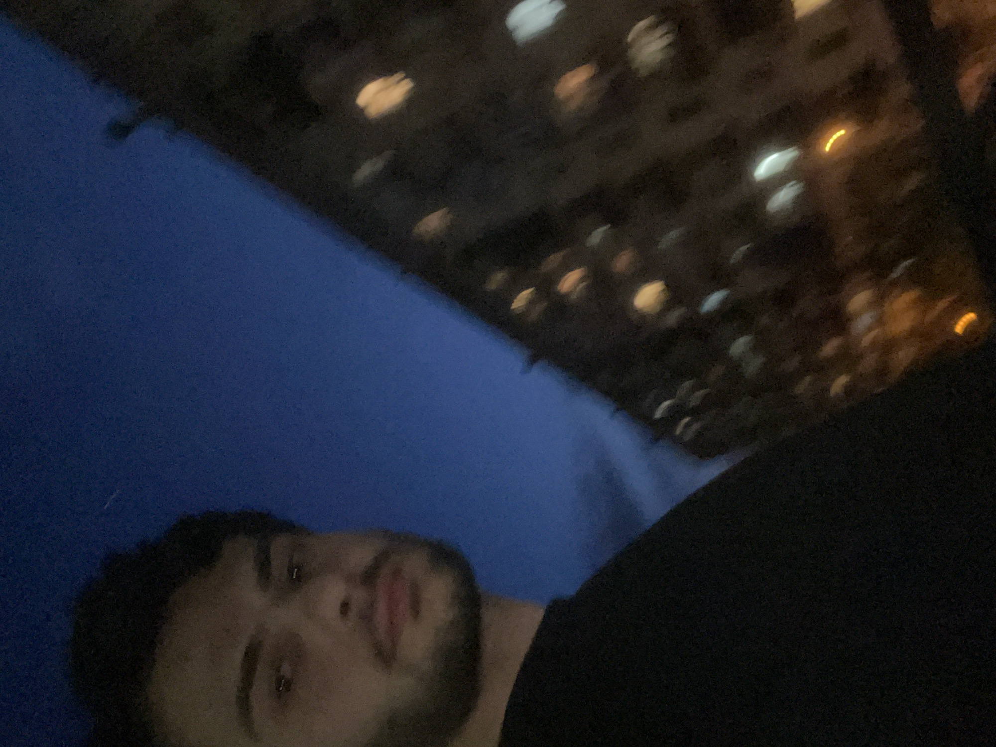 The image shows a man looking up at the sky and the underside of a building at night. The building appears to be located in Tbilisi, Georgia, based on the architectural style and urban context suggested by the blurry lights. The man has dark hair and a beard. He is wearing a dark shirt. The sky is a deep blue color. The underside of the building is dark, with blurry spots of light that could be windows.