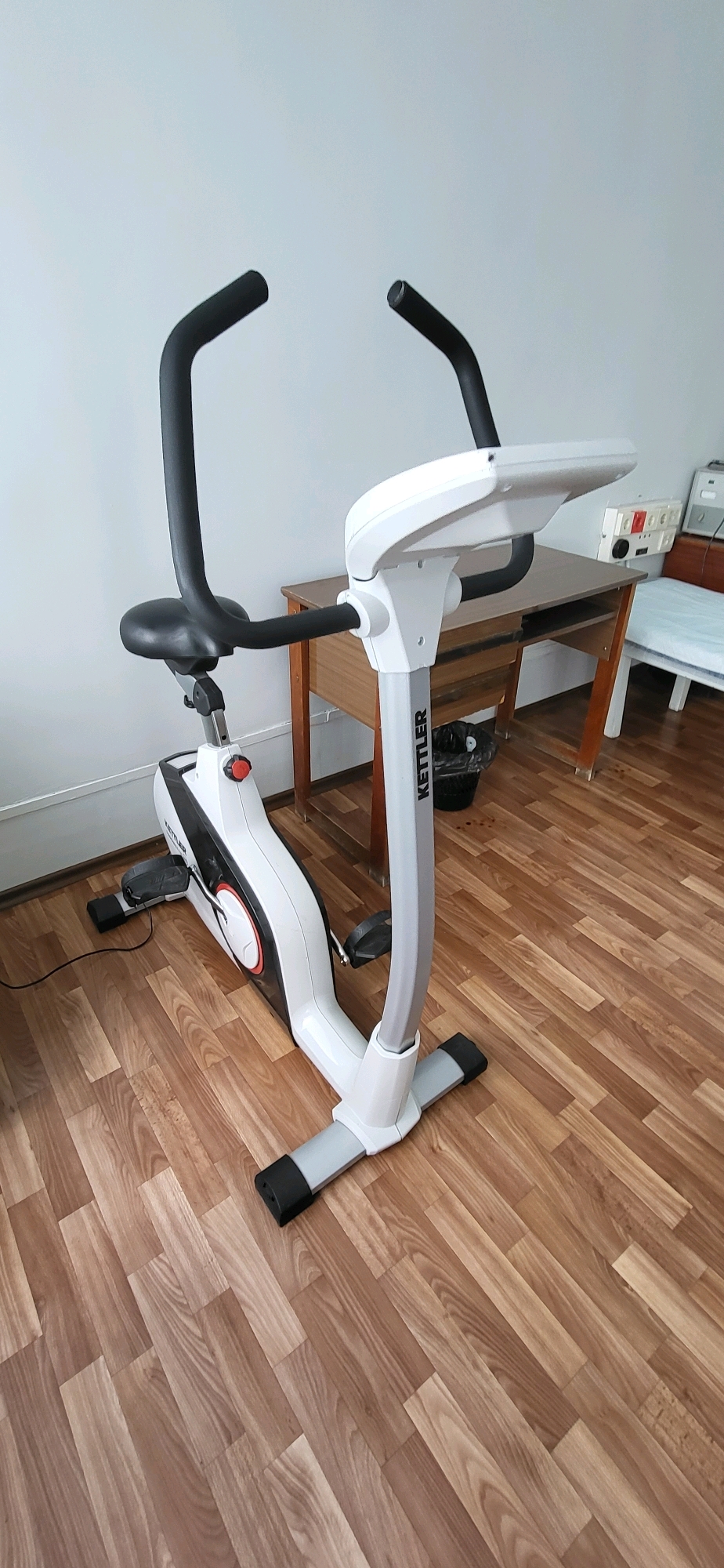 An indoor shot shows a Kettler exercise bike on a wooden floor in a room. The bike is white and gray with black handlebars and seat. A desk and a trash can are visible in the background, along with a chair with a blue cushion. A power outlet is mounted on the wall. The scene is likely a residential setting in Oskemen, Kazakhstan.