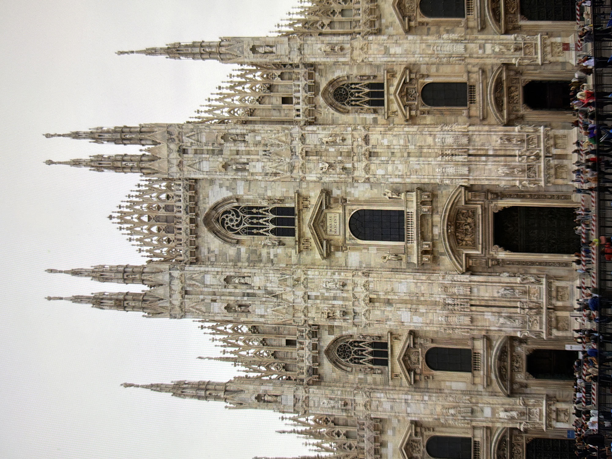 This is an image of the Milan Cathedral, also known as the Duomo di Milano, located in Milan, Italy. The image shows the facade of the cathedral, characterized by its Gothic architecture, spires, intricate carvings, and numerous statues. The sky is overcast. In the foreground, a crowd of people is gathered, some standing behind a metal barrier. The scene depicts a public space in front of a prominent historical and religious landmark.