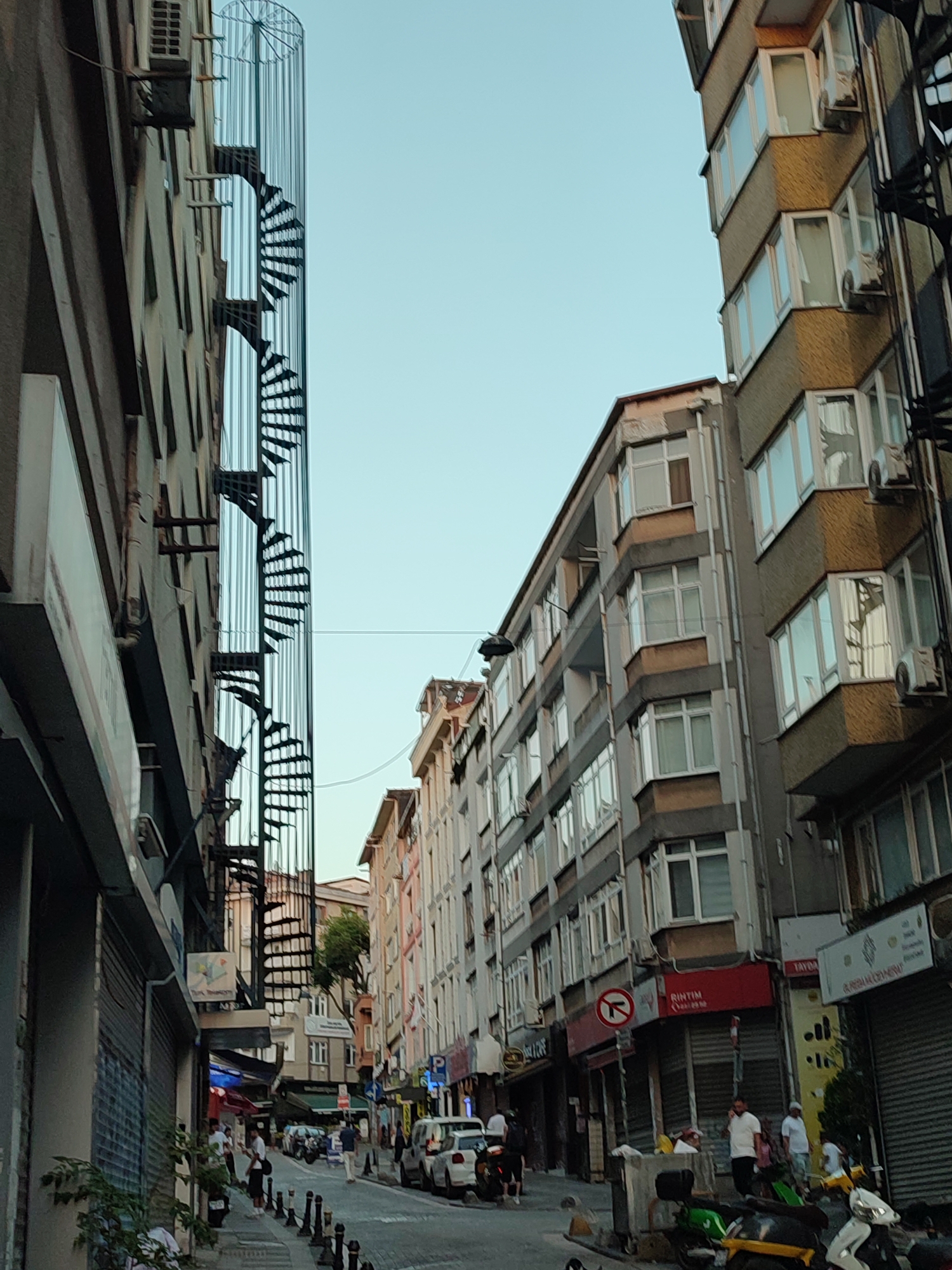 The image depicts a street scene in Kadıköy, Turkey, characterized by a narrow street flanked by multi-story buildings. The architecture is predominantly modern, with balconies and numerous windows. A prominent feature is a metal spiral staircase attached to the exterior of one building, rising vertically along its facade. The street itself is paved and appears to be moderately busy, with several parked cars and motorcycles. A few individuals are visible, some walking and others standing, suggesting a pedestrian-friendly environment. Commercial establishments, such as shops with rolling shutters, are present along the street level. The overall impression is of a lively urban setting, likely a commercial or residential area within the city. The sky is clear and blue, indicating daytime.