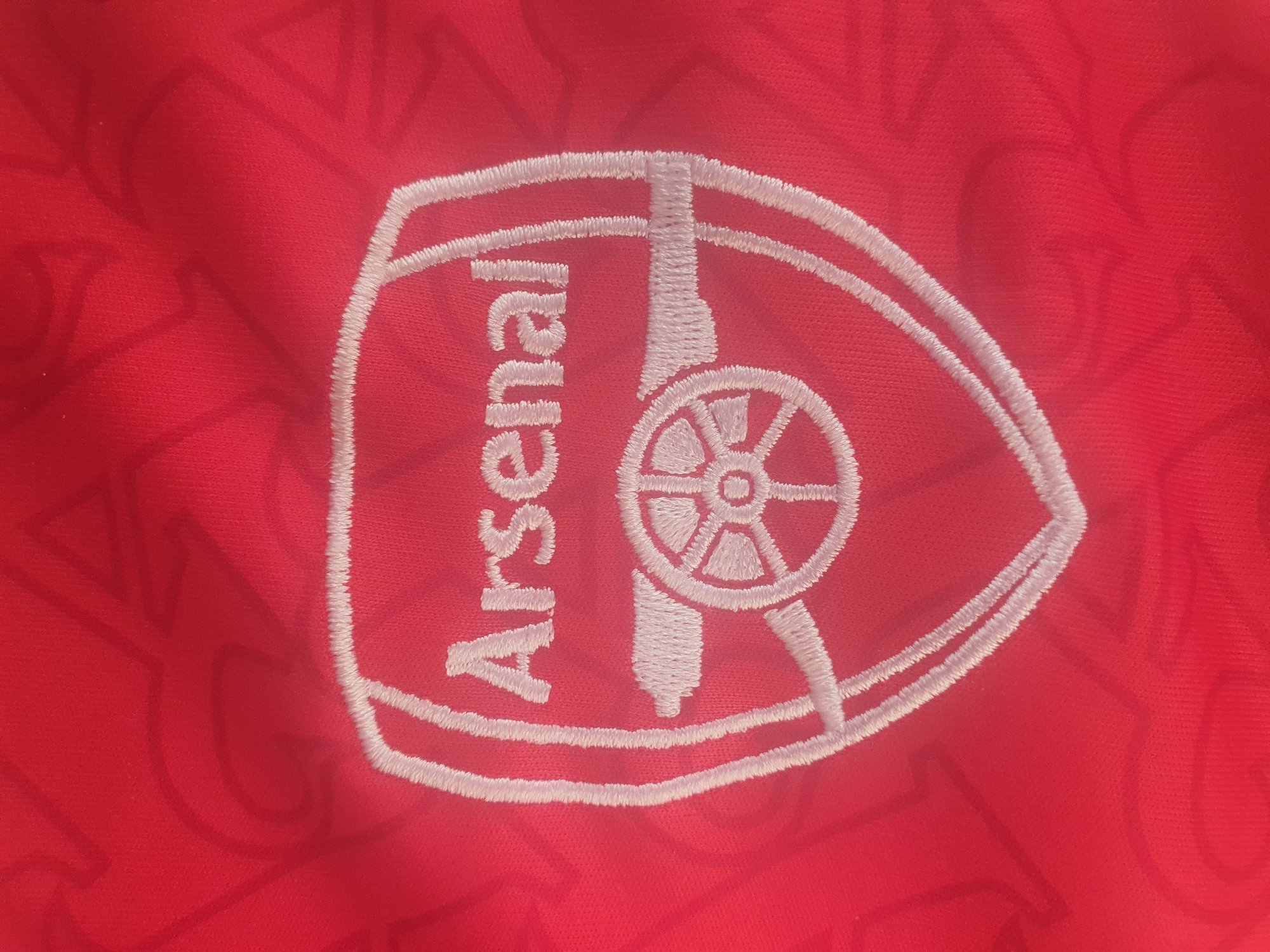 The image is a close-up shot of an embroidered Arsenal Football Club crest on a red fabric, likely a jersey or scarf. The crest is white and features the club's name "Arsenal" arched above a cannon. The fabric itself has a subtle, lighter red pattern embossed on it, possibly a watermark or design feature of the material. The lighting is even, highlighting the texture of the embroidery and the weave of the fabric. There are no people or discernible environmental details present in the image. The location of Jos, Nigeria, is not visually represented in this image.