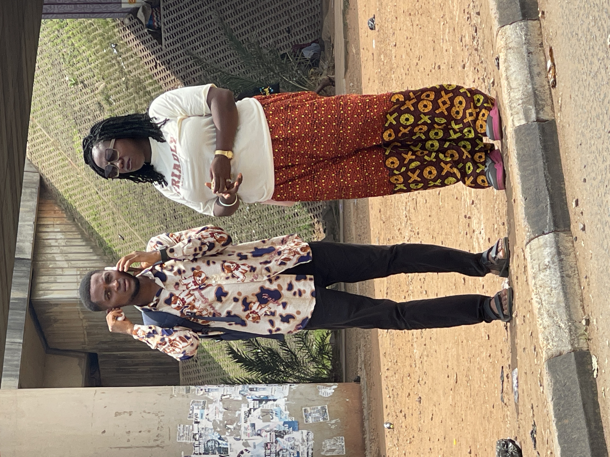 Two people stand on a street, likely in Abuja, Nigeria. A woman in a white t-shirt and patterned orange and brown pants stands to the right, wearing sunglasses and a watch. A man in a patterned shirt and black pants stands next to her, holding a phone to his ear. They are on a sidewalk next to a street with a concrete curb. A wall covered with posters is visible in the background.