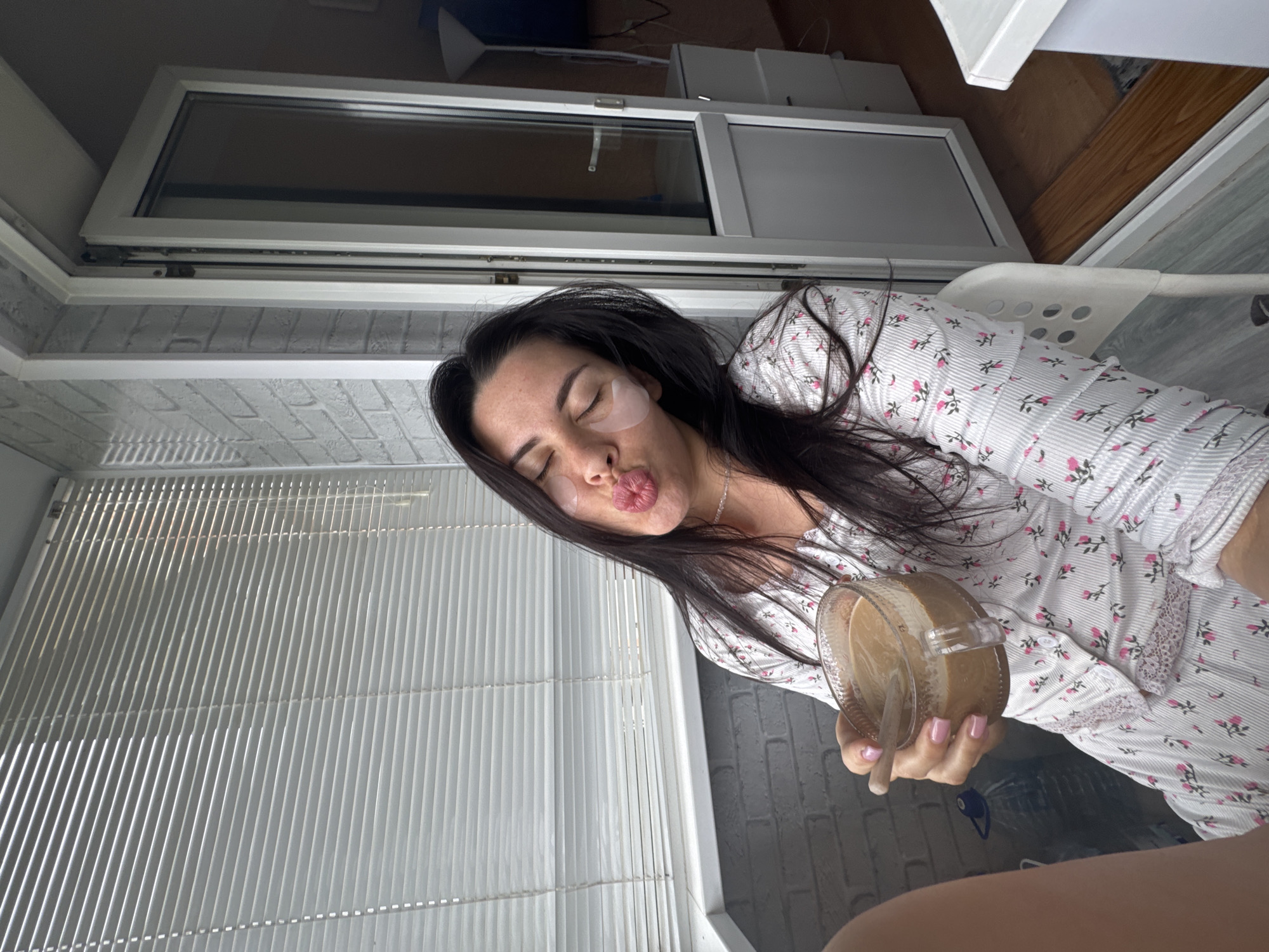 A woman with dark hair and eye patches is sitting on a white chair, holding a clear glass mug of coffee with a wooden spoon. She is wearing a white pajama set with a floral pattern. She is puckering her lips and has her eyes closed. The background includes blinds on a window, a brick wall, and a glass door. The image is taken in an indoor setting, possibly a balcony or room with a view of the outdoors. There is no visible indication of the city of Chaikovsky, Russia.