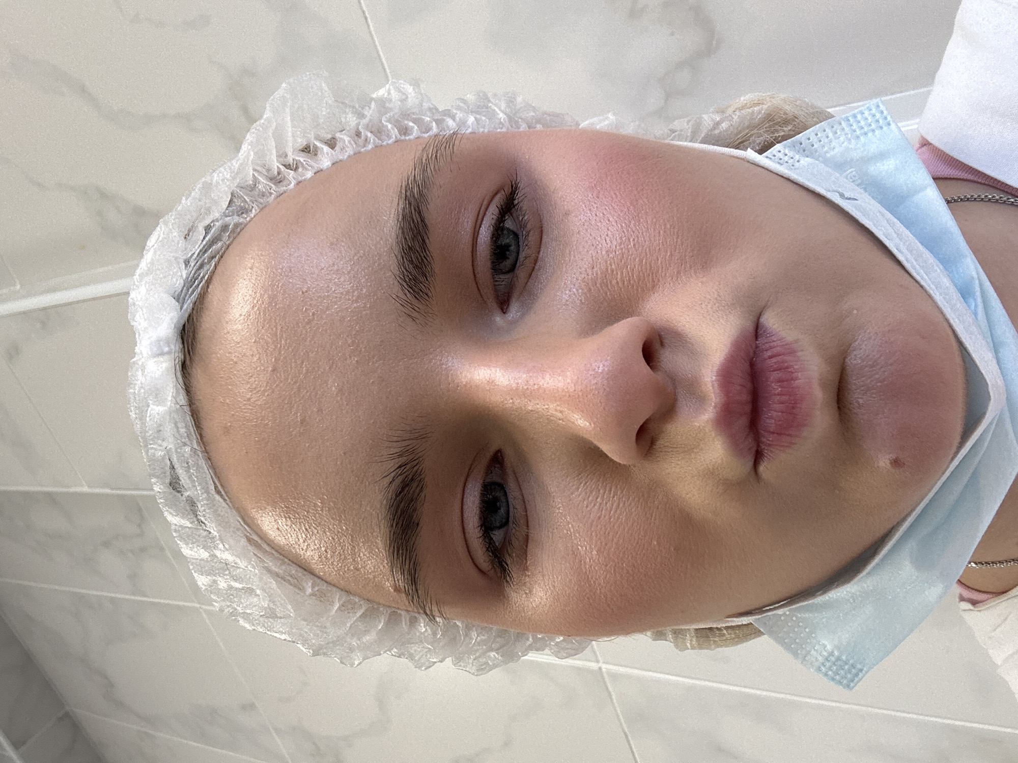 A fair-skinned woman lies reclined with a medical hair net and a blue medical face mask. The backdrop is white with some faint marble-like patterning, likely a tiled wall or floor. No identifiable location in Saint Petersburg, Russia is discernable based on the provided image.