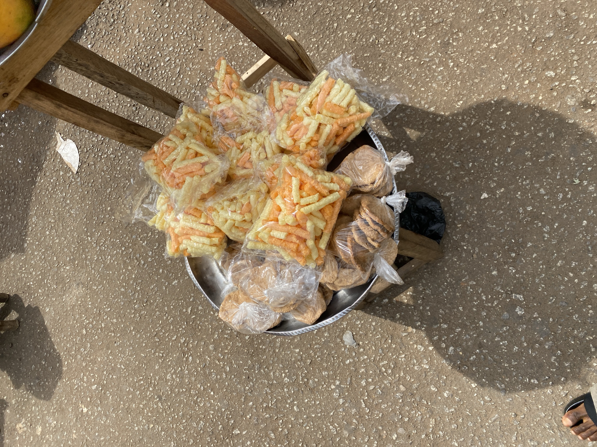 The image, captured from an overhead perspective, depicts an assortment of individually packaged snacks displayed in a large metallic tray on a textured, light-colored ground, consistent with asphalt or concrete in an outdoor setting like a market or street in Bukuru, Nigeria.
The tray is filled with numerous clear plastic bags containing two distinct types of snacks. On the left and upper sections, there are many bags filled with light yellow and orange-colored, cylindrical or stick-shaped crisps or puffs. On the right and lower sections, other plastic bags hold flat, round, brown-colored items, resembling cookies or crackers. A black plastic bag is partially visible on the right side of the tray.
Part of a wooden structure, likely a stool or table leg, is visible in the upper-left quadrant, with a hint of a yellow-orange fruit in another metallic container just beyond it. Strong, defined shadows are cast on the ground from the wooden structure, the tray, and a person standing nearby, indicating bright sunlight and clear, daytime weather. A human foot, wearing a black sandal, is visible in the bottom-right corner of the frame. The scene suggests a street vending operation where snacks are offered for sale. No visible text is present on any of the items.
