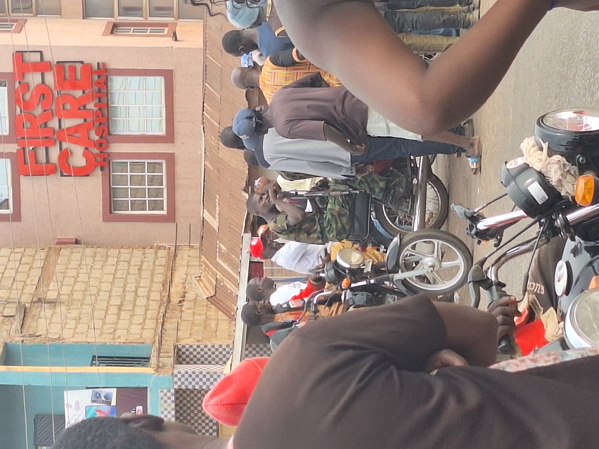 In Sabon Tasha, Nigeria, an outdoor scene shows several people standing near motorcycles on a street in front of a building bearing the name "First Care Hospital" in red letters. The building has multiple windows. Individuals are dressed in various attire, including casual wear and military-style clothing. Some are seated on the motorcycles. The surroundings include portions of the street and elements of the urban environment.