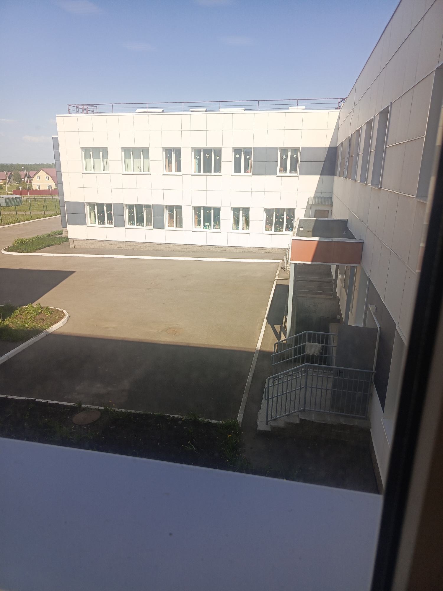 The image captures an exterior view of a modern, two-story building with a predominantly white facade, likely located in a suburban area within the city of Unknown, Unknown. The building has a flat roof and is constructed with rectangular panels. The facade features numerous windows with white frames.
A concrete courtyard occupies the foreground, with visible shadows cast across its surface. There's a set of stairs with a metal railing leading up to a covered entrance area. A small patch of greenery with some vegetation is visible at the edge of the courtyard.
In the background, a distant residential structure is discernible, along with a fence and additional greenery. The sky above is clear and blue, indicating a sunny day. The image appears to have been taken from an elevated position, possibly from within another building looking outward. There are no people visible in the frame. The overall impression is that of a quiet, possibly educational or administrative, setting.