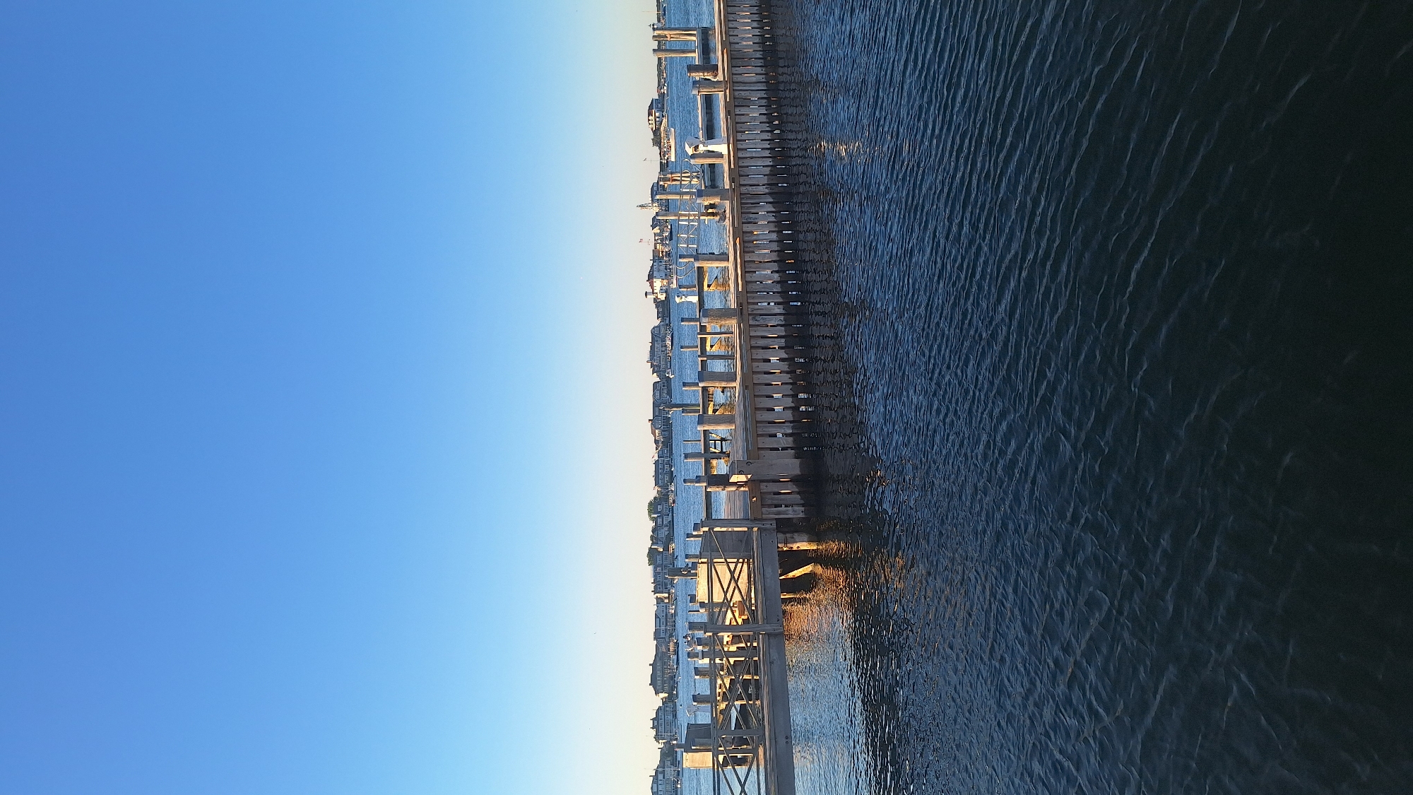 A wooden pier extends into a body of calm, dark blue water under a clear, bright blue sky. The pier is made of weathered wood with vertical planks forming a railing. Several posts support the pier, and some white structures, possibly boat lifts or bollards, are visible on its surface. In the background, a row of houses with light-colored roofs lines the distant shore, and several boats are moored in the water. The scene appears to be a waterfront area in Nantucket, Massachusetts, USA, bathed in the warm light of late afternoon or early morning, casting gentle reflections on the water. There are no people or animals visible in the image.