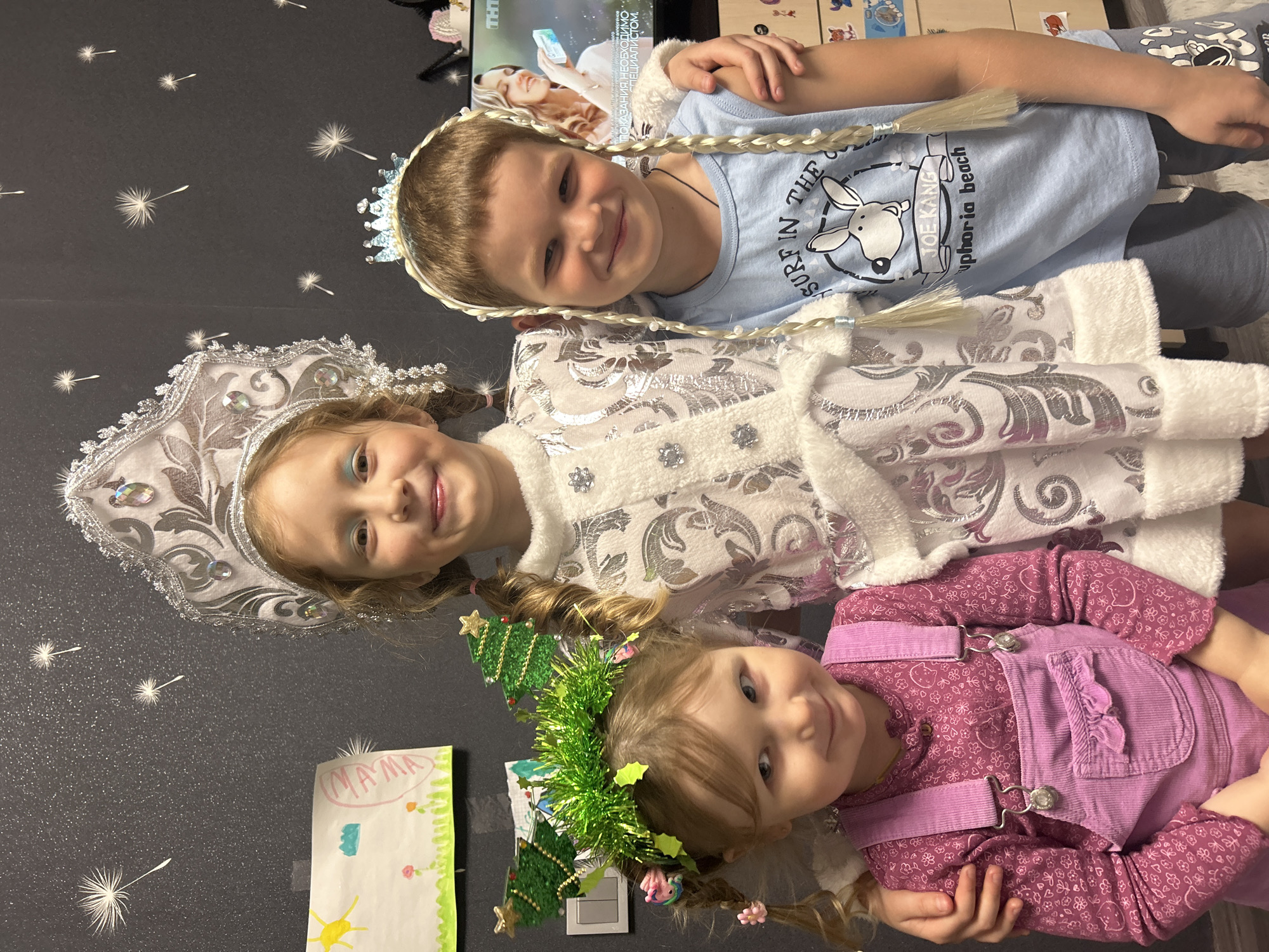 Three children are lying side-by-side. The first child wears a silver and white dress with a matching crown, and the second child wears a light blue shirt and a crown. The third child wears a pink outfit with a green and gold Christmas tree crown. A drawing with the word "MAMA" on it is to the left of the bottom child. The TV in the background is broadcasting a show, and there's text that includes "ПОКАЗАНИЯ. НЕОБХОДИМО СПЕЦИАЛИСТОМ." Another line of text has the words "SURF IN THE JOE KANG uphoria beach." The wallpaper appears to be dark with white, dandelion-like designs.
