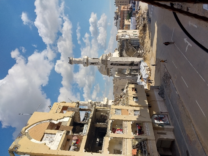 The image depicts a devastated urban landscape in Khan Yunis, Palestinian Territory, under a partially cloudy blue sky. In the foreground, a multi-story building, likely residential, stands heavily damaged on the left, with significant structural collapse revealing interior rooms. Laundry hangs on balconies. Bullet marks and craters are visible on its facade, and debris piles at its base. A paved street with white lane markings, including turn arrows, runs across the lower portion of the scene.
In the midground, a large mosque features a tall, slender minaret and a dome, both showing visible damage. Extensive rubble surrounds the mosque, indicating severe structural failure of adjacent sections. Several individuals are present: one person in a yellow top walks right on the street, another in a dark top stands centrally, and a small group moves amidst the debris near multiple white tents in the open area between the street and the mosque. Further buildings, some appearing damaged, extend into the distance on the right. The overall scene is characterized by widespread destruction and scattered building materials.