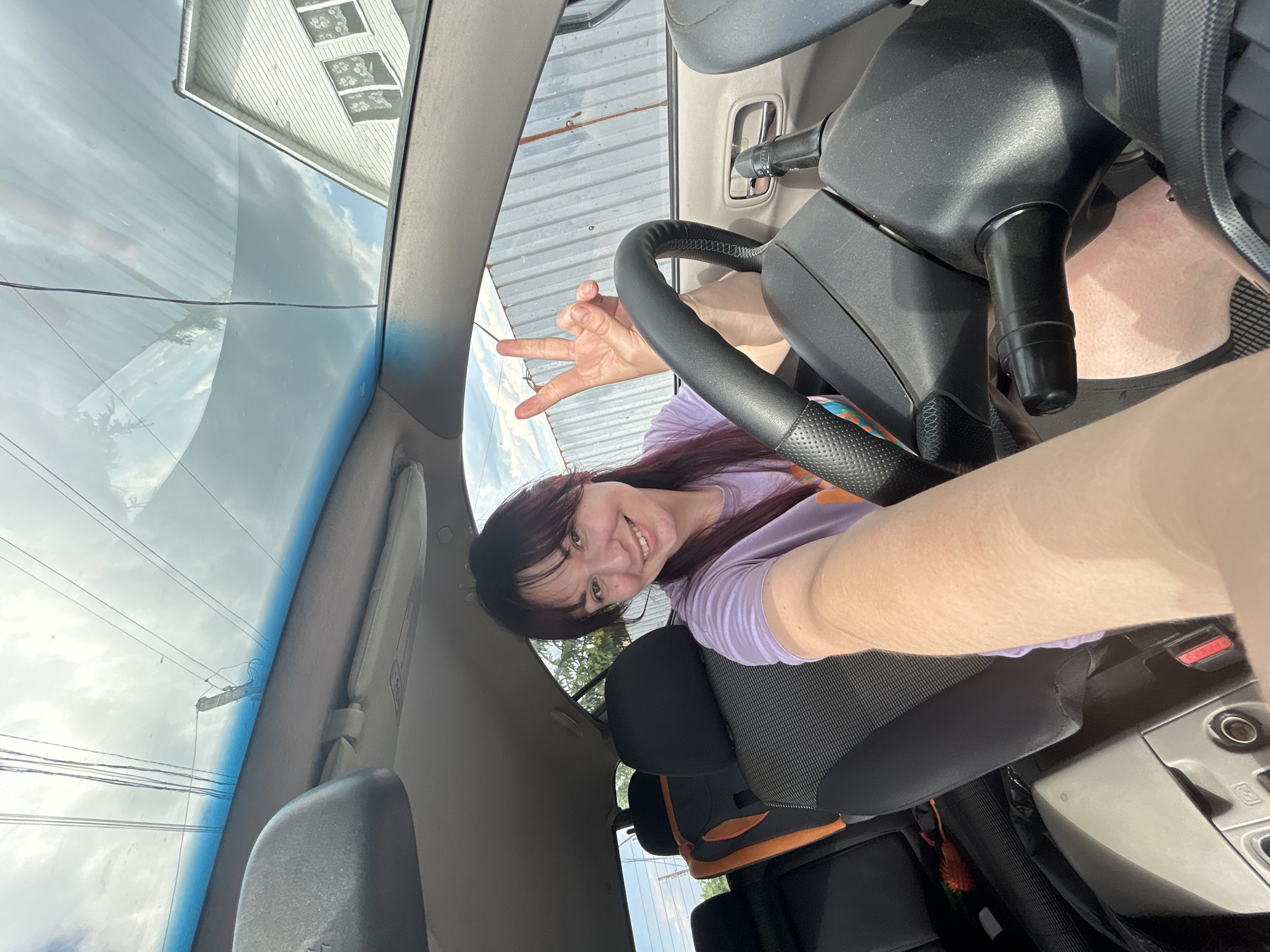 The image shows a young woman with dark hair and bangs, wearing a purple t-shirt, sitting in the driver's seat of a car. She is smiling and holding up two fingers in a peace sign. The car appears to be parked, with the steering wheel and dashboard visible. The interior of the car includes car seats in the back. Outside the car, a white building with windows and a corrugated metal fence can be seen. The sky is cloudy with visible power lines. The photo was taken during the daytime, as indicated by the lighting. Based on the provided information about the city, the scene is located in Фоки, Russia.