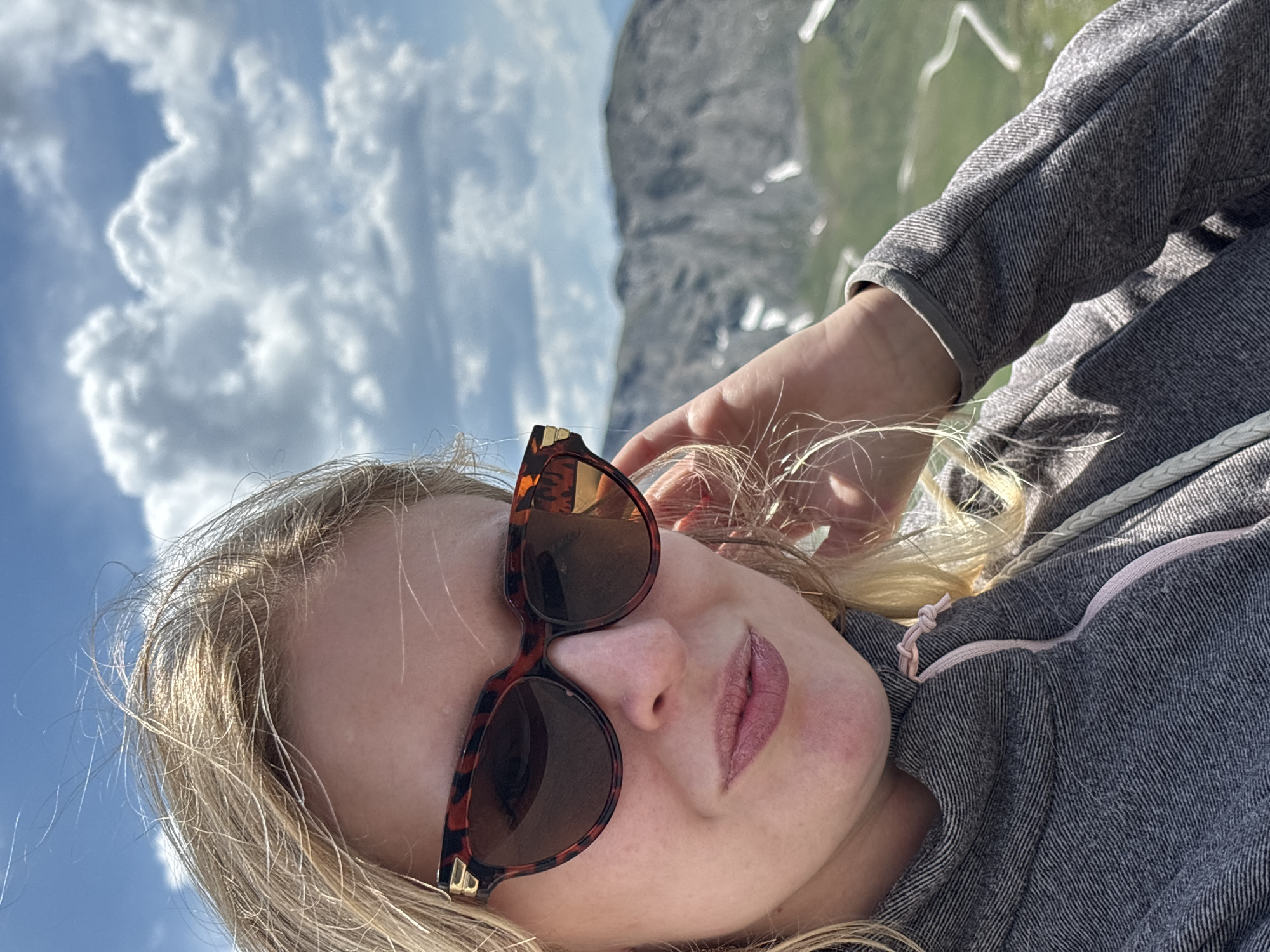 The image shows a woman wearing sunglasses and a gray hooded jacket, taking a selfie outdoors. She has blonde hair and is wearing pink lipstick. The background features a blue sky with white clouds and a mountainous landscape. This scene is located in the municipality of Heiligenblut am Großglockner, Austria, known for its alpine environment. The woman is holding her hand up to her hair. The jacket has a pink drawstring.