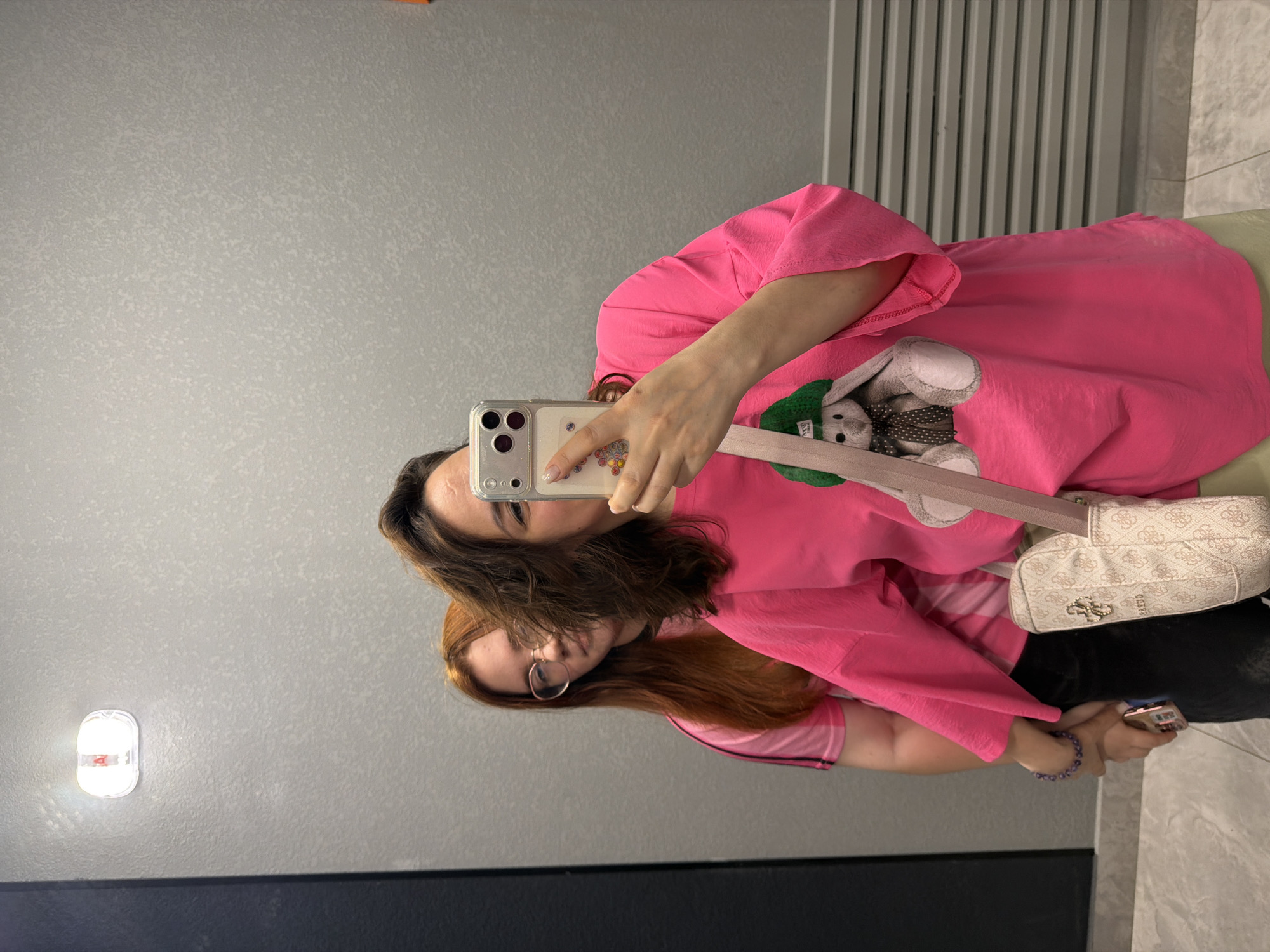 Two young women are standing in front of a grey, textured wall, possibly in a public restroom or changing room. The woman in the foreground is taking a mirror selfie with a smartphone. She has dark, medium-length hair and is wearing a bright pink oversized t-shirt with a graphic of a stuffed bunny wearing a green hat and scarf. A beige crossbody bag with a repeating logo pattern is slung over her shoulder. Her left arm is extended, holding the phone, and her right arm is bent.
The second woman, standing behind and to the left of the first, has long, reddish-brown hair and is wearing glasses. She is looking towards the camera with a neutral expression. She is wearing a pink t-shirt with darker pink stripes on the sleeves. Her left hand is visible, holding a phone and a beaded bracelet.
The environment appears to be indoors, with a ceiling light fixture visible at the top of the frame and a grey, ribbed ventilation grate on the right side of the wall. The floor is a light-colored, textured tile. The overall lighting is somewhat dim. The image has a casual, candid feel. The location is the restroom of a store in the city of Perm, Russia.