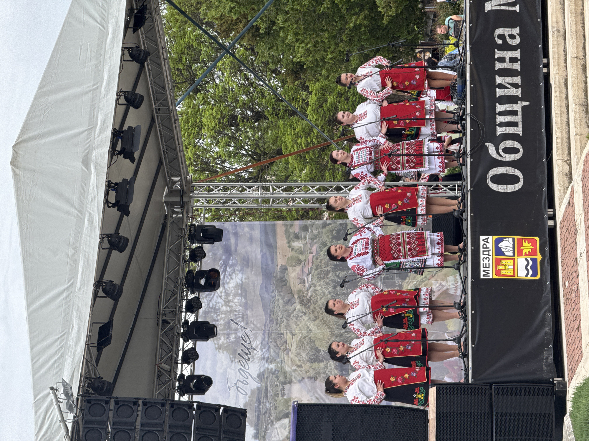 A horizontal, eye-level shot shows a group of women standing in a row on a stage. The women are wearing white tops and red and black skirts with embroidery. Each woman has a microphone in front of her. Behind the women is a backdrop with a scenic view. To the left of the women is the stage lighting and structure. To the right of the women is a black banner with the word "Община" written in large white letters. Below that is the coat of arms for the city of Mezdra, Bulgaria. The banner also says "МЕЗДРА." The stage is located in an outdoor area, and trees are visible in the background. The ground is paved with stone. The lighting on the stage suggests a daytime performance.