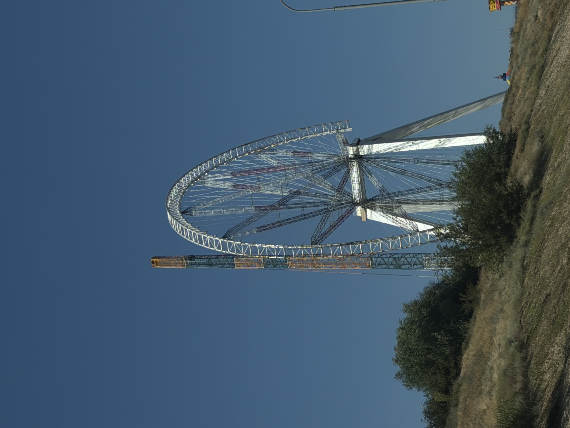 The image depicts a large Ferris wheel, likely under construction or undergoing maintenance, in Volgograd, Russia. The Ferris wheel is silver and white with red accents on some of its spokes. To the left of the Ferris wheel, a tall, narrow, orange and blue structure, possibly a construction crane or a support tower, stands against a clear blue sky. The Ferris wheel and the tower are situated on a grassy, hilly terrain with scattered bushes and trees. In the distance, on the right side of the image, a street lamp is visible. The overall scene is a daytime outdoor shot, focused on the industrial or amusement park structure.