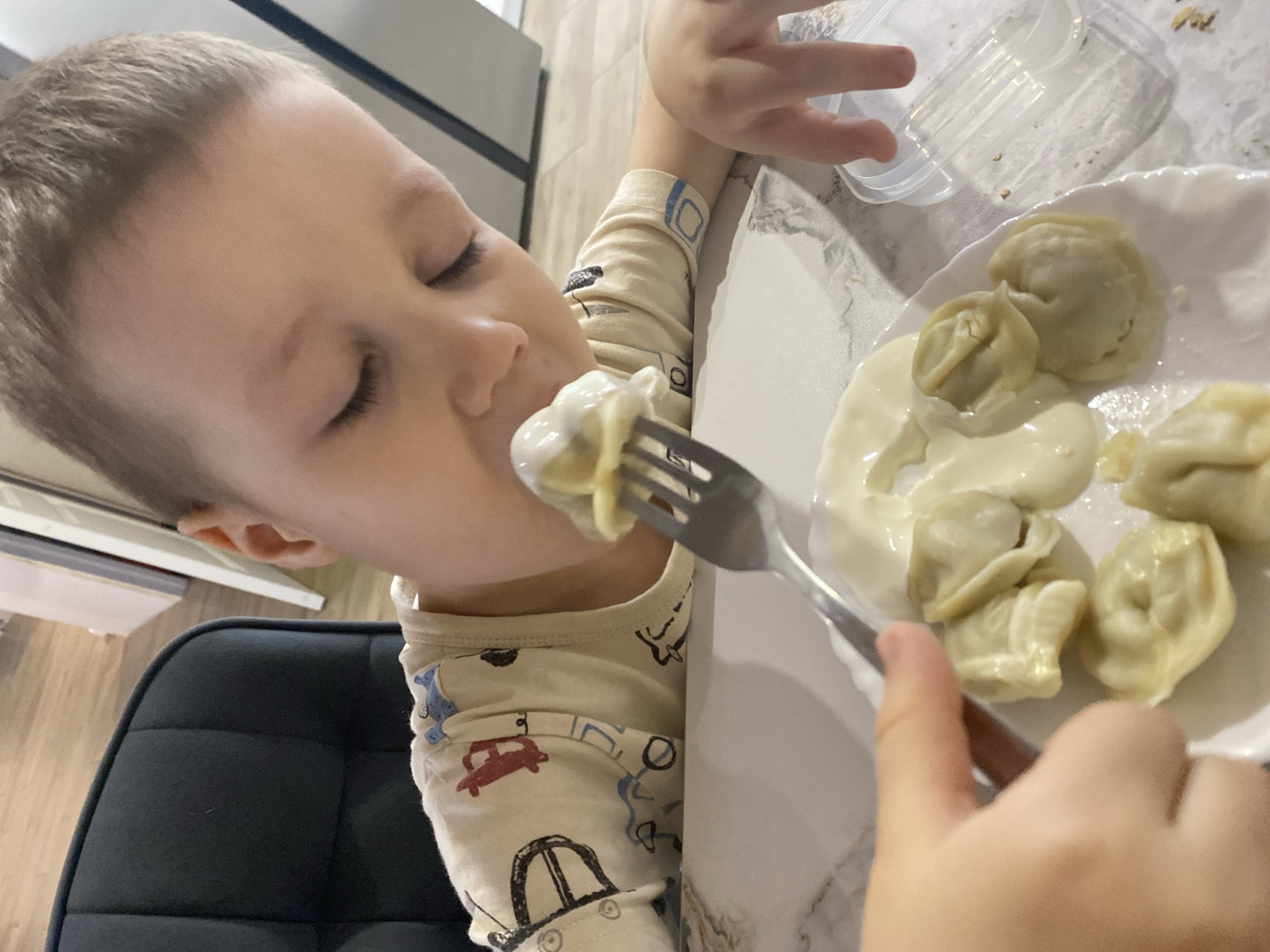 A young child is captured mid-meal in what appears to be a brightly lit indoor setting, likely a kitchen or dining area. The child, with light-colored hair, is positioned on the left side of the frame, facing slightly right, with eyes gently closed. They are wearing a light-colored long-sleeved shirt or pajamas adorned with various car patterns.
The primary activity depicted is the child eating. A silver fork holds a dumpling, which is covered in a white, creamy sauce, and is positioned near the child's mouth. The child's right hand holds the fork. On a white, scalloped-edge plate, several other similar dumplings are visible, also generously coated in the white sauce, resembling sour cream or smetana, a common accompaniment for dumplings like pelmeni, consistent with the provided context of Chaikovsky, Russia.
The table surface beneath the plate is a light-colored marble or speckled countertop. In the background, out of focus, parts of a modern interior are visible, including what looks like kitchen cabinetry and wall. A dark, padded chair is partially visible on the bottom left, suggesting the child is seated at a table. The lighting suggests it is daytime. The child's closed eyes could indicate deep concentration or enjoyment of the food. No explicit text is visible in the image.