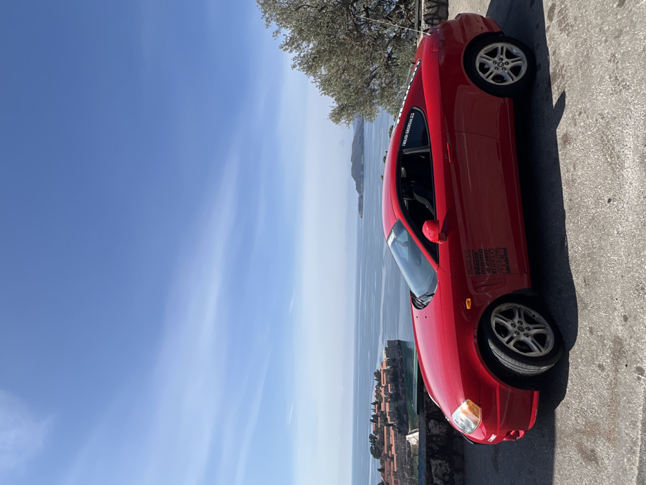 A red two-door coupe, identified as a Hyundai Tiburon based on visible styling cues, is parked on an asphalt surface, facing left in the frame. The vehicle features various sponsor decals on its lower front fender, including "SPARCO," "NRG," "MOMO," "Nardi," "HKS," and "WRC." A sticker displaying "TBILISI GEORGIA" with the Georgian flag is affixed to the passenger-side rear quarter window. The car is equipped with silver five-spoke alloy wheels.
The scene is set in Sveti Stefan, Montenegro, with the fortified island of Sveti Stefan prominently visible in the mid-ground, characterized by dense clusters of red-roofed stone buildings connected to the mainland by a narrow causeway. The calm, blue Adriatic Sea extends behind the island, fading into the horizon where faint landmasses are visible. The sky is clear blue with minimal, wispy white clouds. To the right of the frame, a portion of an olive tree with silvery-green foliage is visible. The foreground shows the paved parking area and a low stone wall separating the viewpoint from the coastline below. No individuals are present in the image. The lighting indicates bright daylight.