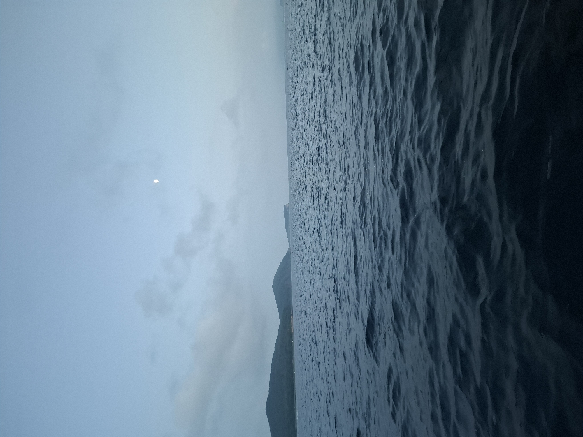 The image shows a vast expanse of dark, choppy water under a pale, overcast sky. In the distance, silhouetted against the sky, are the outlines of rolling hills and mountains. A sliver of moon is visible in the upper right quadrant of the sky. The scene is devoid of any human presence, vessels, or structures. The overall impression is one of solitude and the raw power of nature. This scene is located in the vicinity of Putyatin Island, a coastal settlement in Russia's Primorsky Krai, characterized by its rugged coastline and mountainous terrain meeting the Sea of Japan.