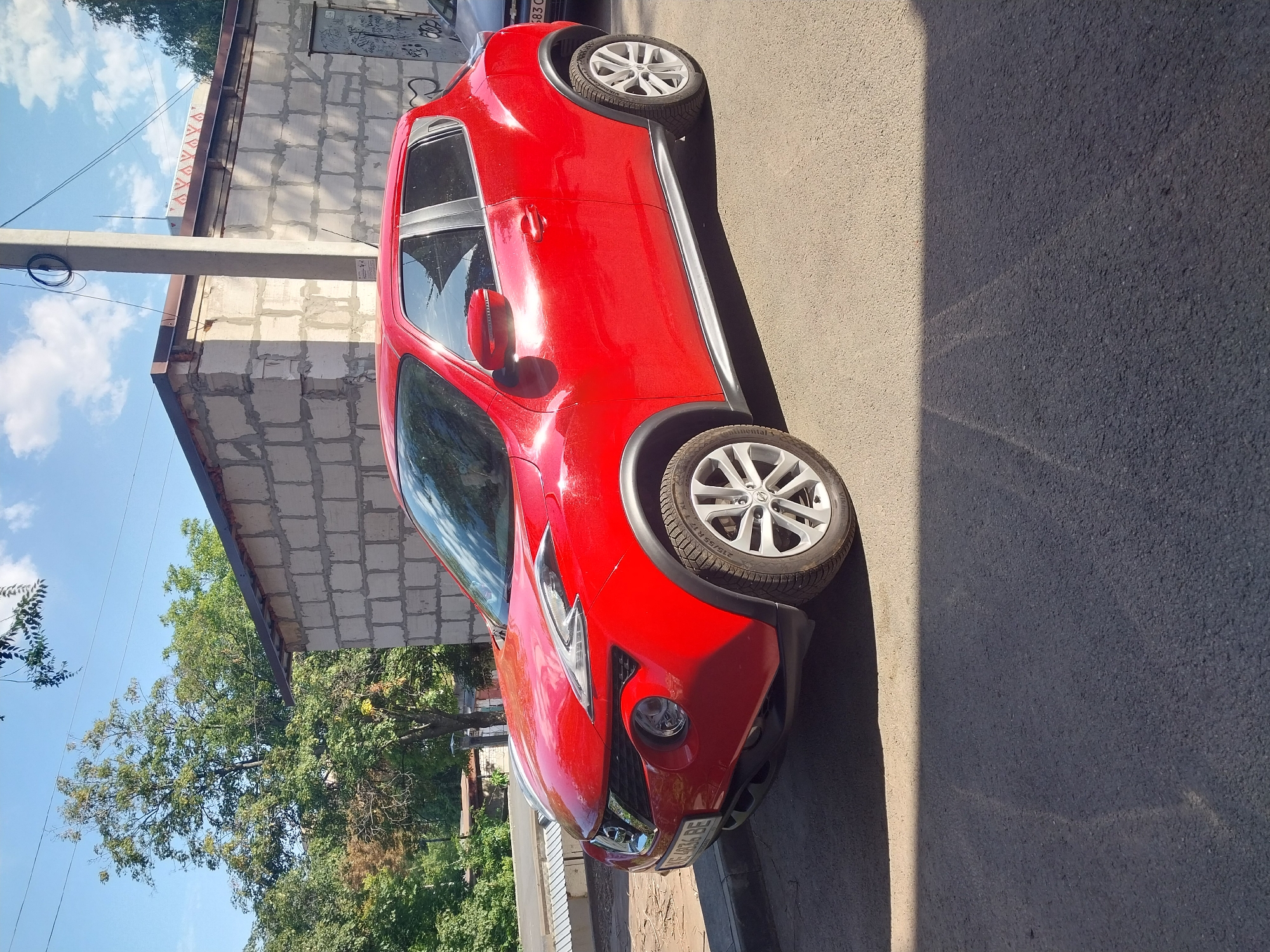 A red Nissan Juke is parked on an asphalt surface next to a building constructed of cinder blocks. The building has a brown roof and a graffiti-covered metal door on the right side. The car's license plate reads "VE 4836 BE". Trees and a utility pole are visible in the background. The sky is blue with scattered clouds. The image was taken in the city of Dnipro, Ukraine.