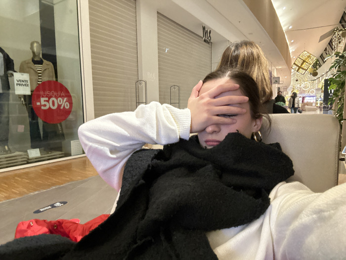A person with dark hair and gold hoop earrings sits in a beige chair, shielding their face with their hand. They are wearing a white sweater and a black scarf. Behind them, another person with brown hair stands. The background shows a shopping mall with closed storefronts, one displaying a "-50%" sale sign. Decorations hang from the ceiling, and people are walking in the distance. Red clothing items are visible on the floor.