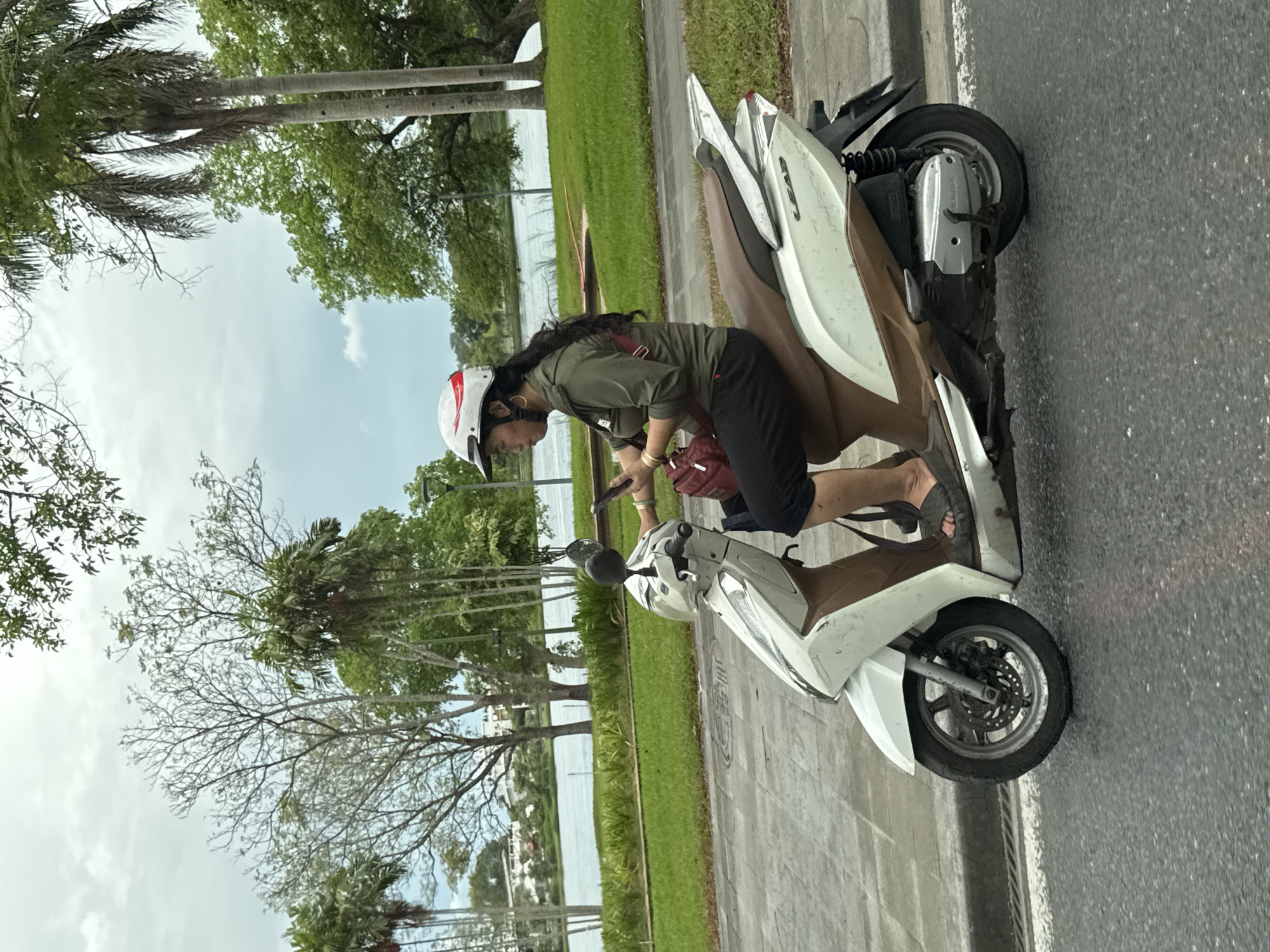 A woman rides a white and brown Honda Lead scooter on a paved road beside a grassy area and a body of water. She is wearing a white helmet with red accents, a green long-sleeved shirt, and black pants. She holds a smartphone in her right hand and a red bag is slung over her left shoulder. Tall palm trees and other foliage line the background, with buildings visible in the distance. The sky is overcast.