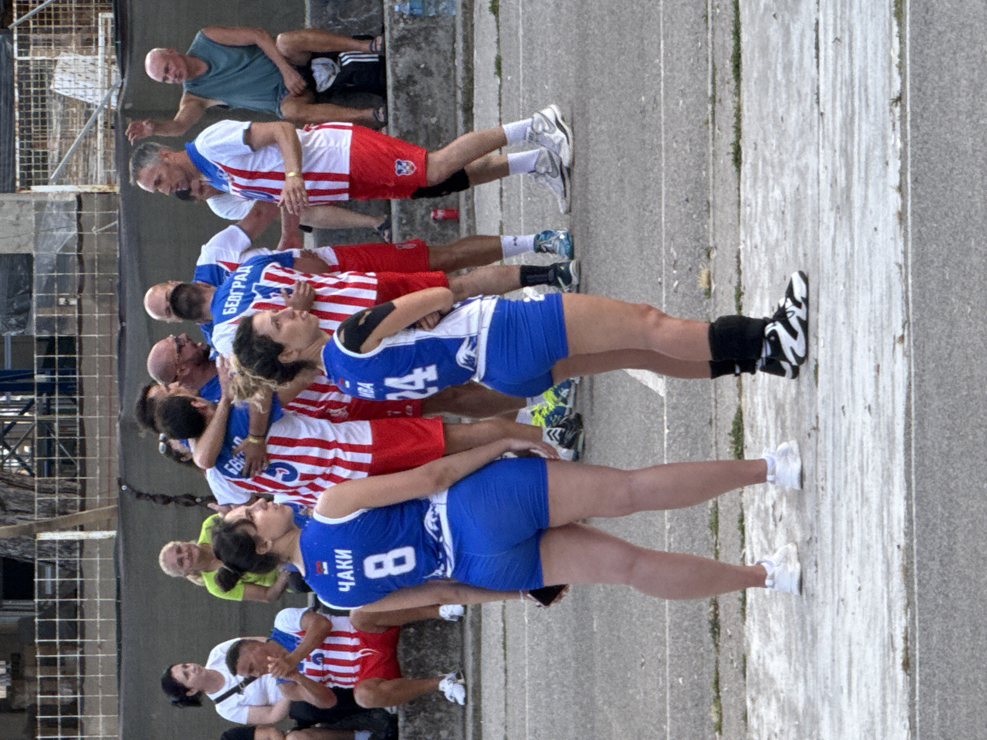 Two women in blue volleyball uniforms stand in the foreground, facing away from the camera. The woman on the left wears a blue tank top with white trim and the number 8 on the back, along with blue shorts. The woman on the right wears a similar blue tank top with white trim and the number 24 on the back, also with blue shorts. Both women are wearing athletic shoes. In the background, a group of people in red and white striped jerseys are gathered, some embracing. A man in a blue and white jersey with red shorts is visible in the middle ground, gesturing with his hands. The setting appears to be an outdoor sports court, with bleachers and a fence in the background. The image is taken in Budva, Montenegro, as indicated by the signage in Cyrillic on some of the jerseys, which reads "BEОГРАД" (Belgrade) and "ЧАКИ" (Čačak), likely indicating the teams' origins or affiliations. The ground is paved, and there is some vegetation visible around the edges of the court.
