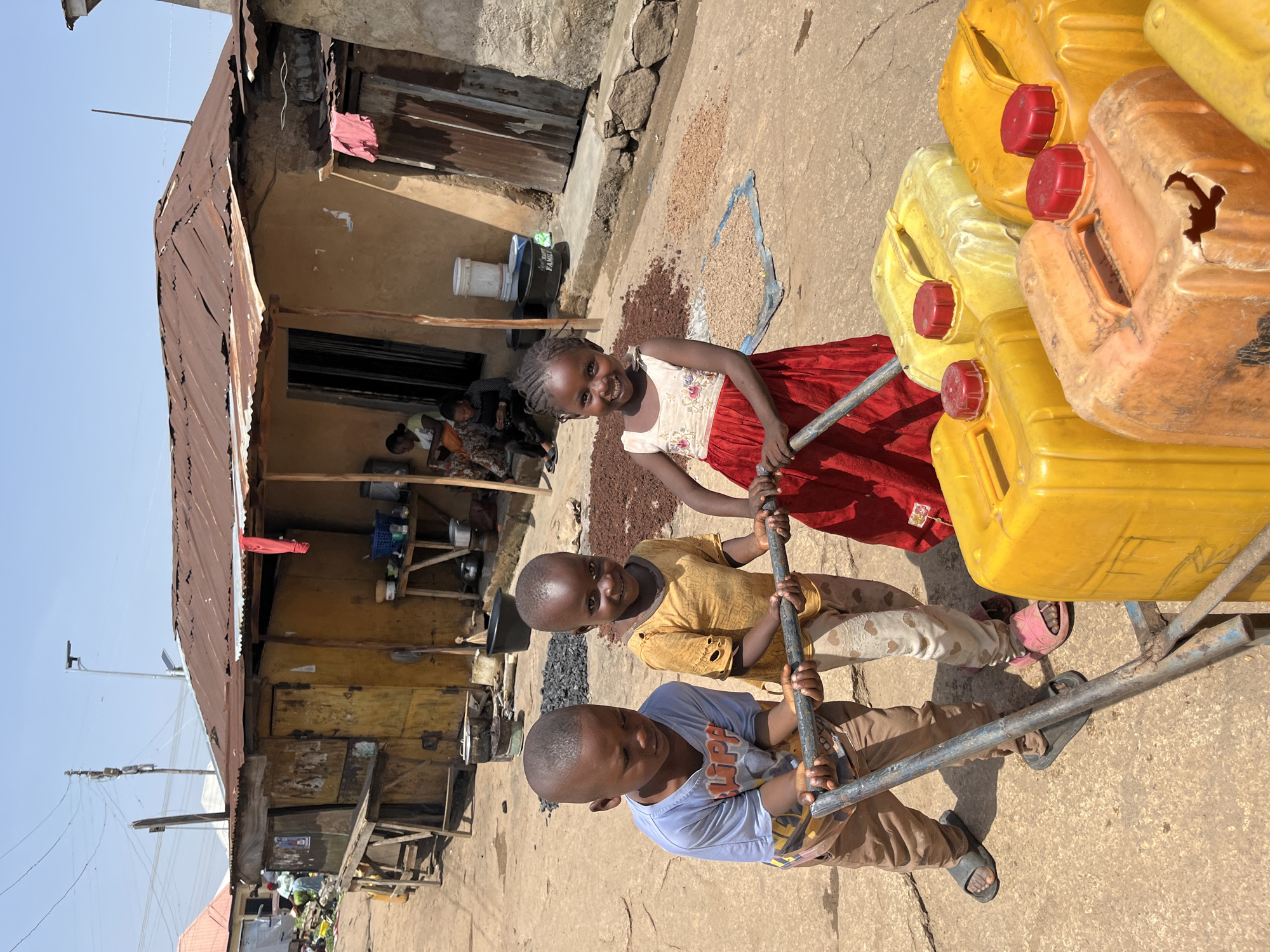 In Jos, Nigeria, three young children are positioned in the foreground, actively engaged in moving several large yellow and orange plastic jerrycans. Two boys, wearing t-shirts and shorts/pants, are pushing a metal cart loaded with the jerrycans. A girl in a red dress is assisting them. Behind the children, a rustic, open-fronted building constructed with mud and corrugated metal roofing stands. Various items, including pots, buckets, and dried goods, are visible both inside and outside the structure. Several piles of dried, brown materials are spread on the ground in front of the building. The environment appears to be an outdoor market or residential area with a dusty, unpaved ground. The sky is clear and blue, suggesting a sunny day.