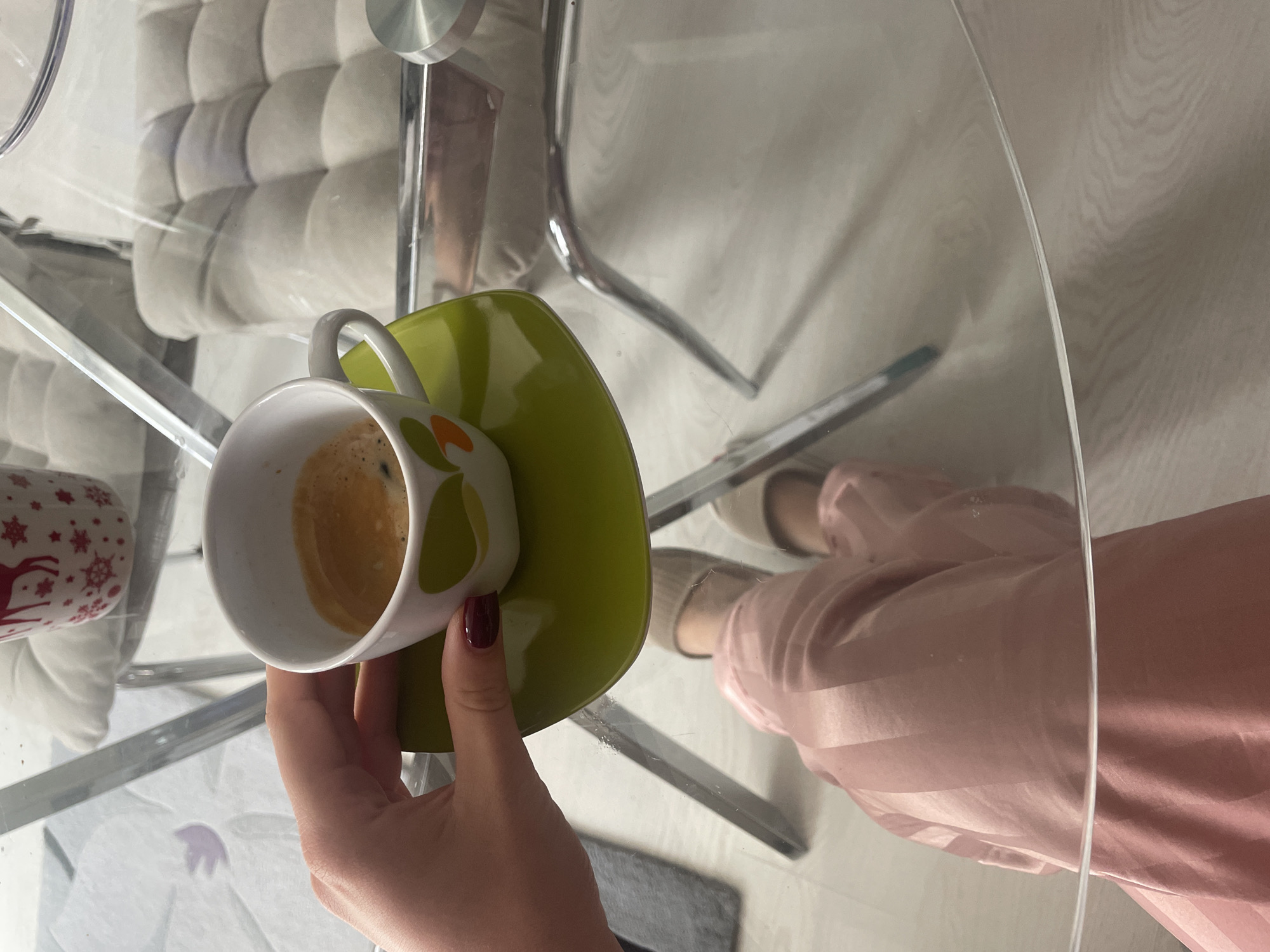 A photograph, apparently taken from directly above, shows a partial view of a glass table with a white and green patterned coffee cup sitting on a matching green saucer in the lower left quadrant. A hand with burgundy nail polish is partially holding the saucer. The coffee cup contains a dark liquid, likely coffee. Beside the coffee cup is another mug decorated with white snowflakes and red reindeer. Part of a white cushioned chair with a chrome frame is visible behind the mugs. Reflected in the glass table are the feet of a person wearing beige slippers and pink striped pants. The environment appears to be indoors, and it is unknown if the location is in the city of Sofia, Bulgaria.
