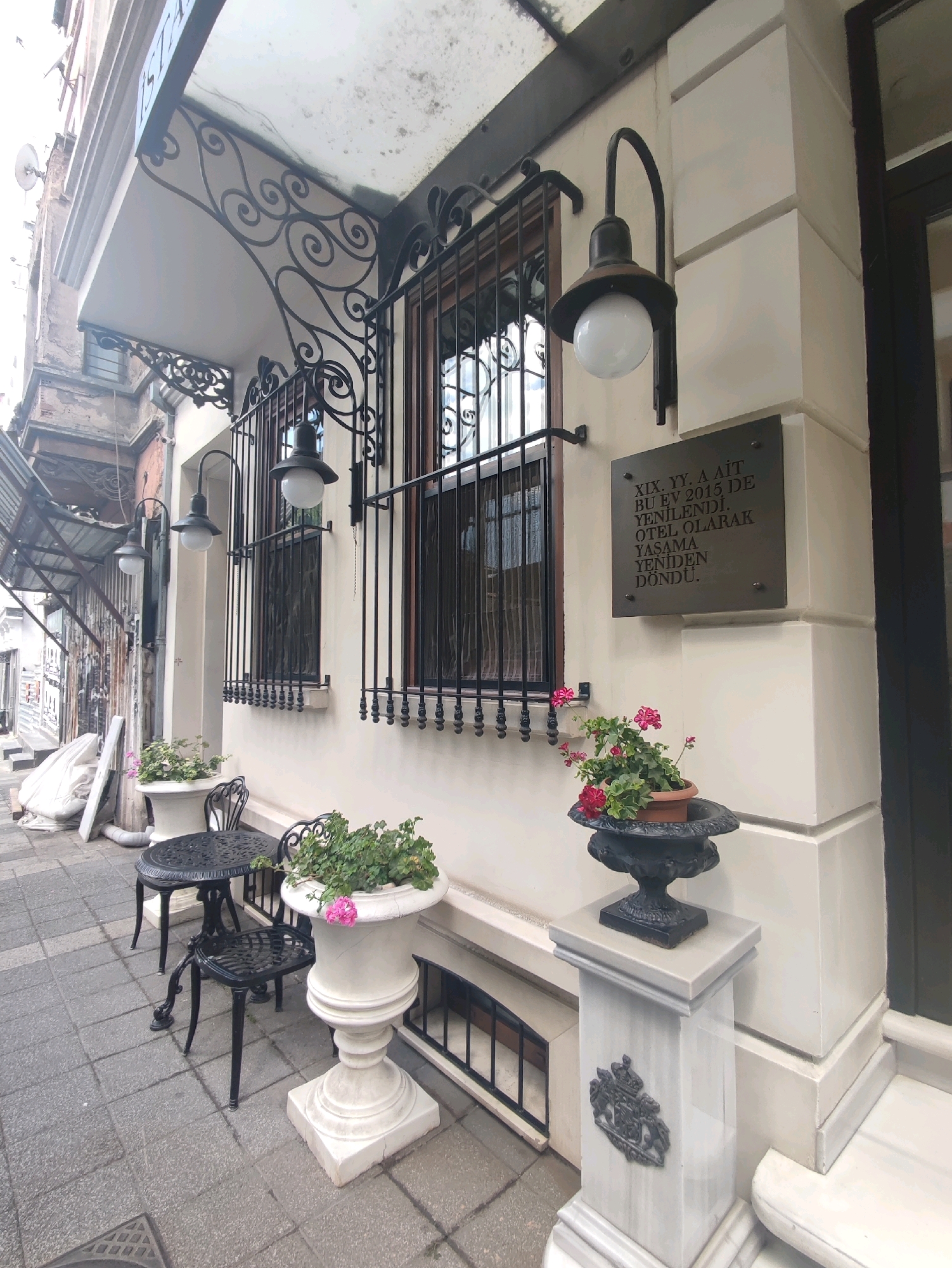 The image captures the exterior of a building in Kadıköy, Turkey, likely a hotel, with a plaque indicating renovations or a re-opening in 2015. The building's facade is cream-colored with decorative wrought-iron window grills and classic outdoor lighting fixtures. In front of the building, a small black metal bistro set with a table and two chairs sits on a gray tiled sidewalk. Adjacent to the table, a large white urn holds a flowering plant. Further to the right, a smaller black planter with pink-flowered plants rests on a marble pedestal that features a decorative crest. The overall scene suggests an inviting and historical urban setting.