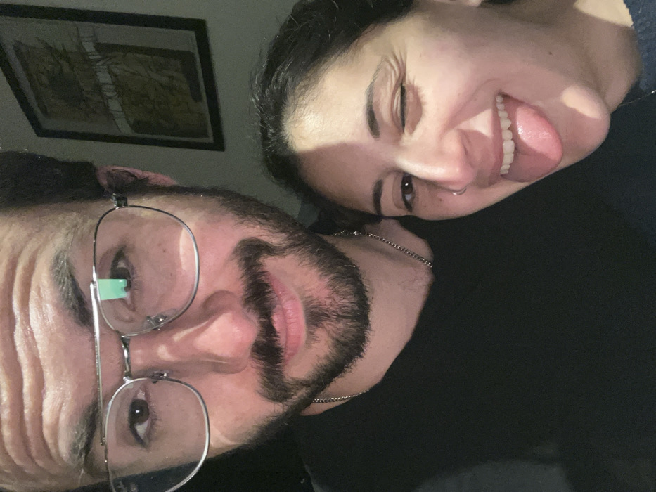 A selfie captures a man and woman in close proximity. The man has dark hair, a beard, and glasses, and he looks directly at the camera. The woman, positioned behind him, winks with her tongue sticking out, creating a playful expression. A framed picture hangs on the wall behind them. The pair is wearing black clothing, and both have dark hair. They both have brown eyes.