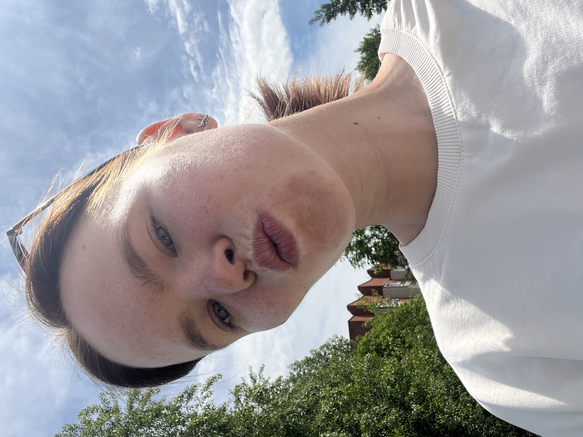A selfie-style photograph shows a person with fair skin, brown hair, and light blue eyes. The person is wearing a white crewneck top. The camera angle is tilted upwards. The background includes a blue sky with white clouds, green trees, and a building with a red roof. The building suggests a setting in Chaikovsky, Russia.