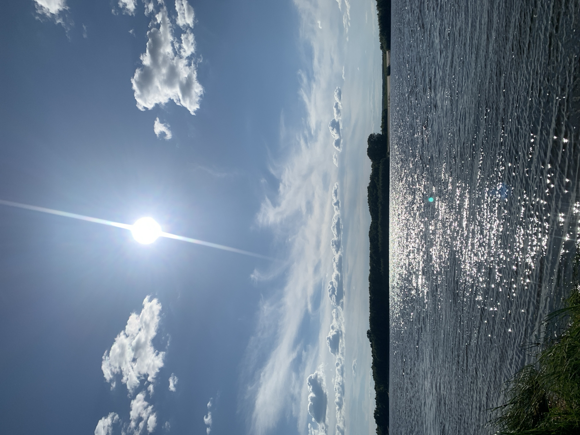 This is a photograph of a scenic natural landscape. The image is oriented vertically, depicting a body of water on the right side and a bright blue sky with scattered clouds on the left. The water's surface is rippling and reflects the sunlight, creating numerous sparkling highlights. A dark line of trees forms the distant shoreline. The sun is visible in the upper left quadrant of the sky, appearing as a bright orb with rays extending outwards. The sky is a clear blue with wispy white clouds and some larger, fluffier clouds. The overall impression is one of a sunny day, likely in the late morning or early afternoon, given the brightness of the sun and the clear skies. The scene evokes a sense of peace and natural beauty. There is no visible text in the image.