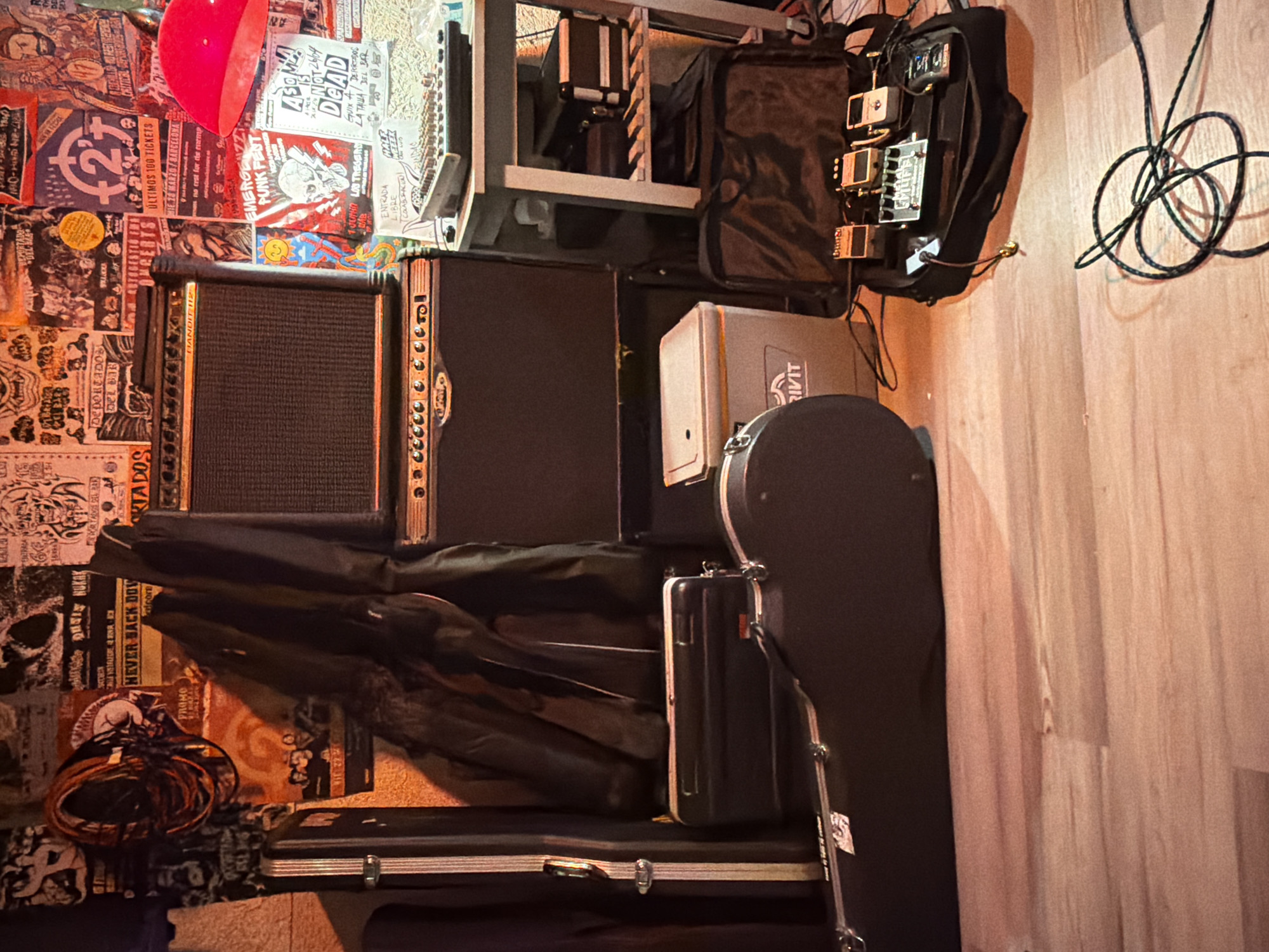 The image depicts a dimly lit room, likely a music practice space or venue, in Sant Adrià de Besòs, Spain. The walls are covered with a collage of posters, predominantly featuring concert flyers and band artwork, many with a punk or hardcore aesthetic. The posters are varied in color and design, with some appearing aged and torn.
In the foreground, a light-colored wooden floor is visible. To the left, a large, dark-colored guitar case lies open. Above and behind the guitar case, a smaller, black case is positioned. Further back, a grey and black cooler with the brand name "RIVIT" is visible.
Dominating the center and right of the frame are two stacked musical amplifiers, black with silver accents. Cables are present, including a coiled black cable on the floor and some running between the equipment.
To the right of the amplifiers, a white metal shelf unit holds various items. On the top shelf sits a mixing board with numerous knobs and faders. Below, on the second shelf, are at least two black cases, one with silver latches. A black bag, possibly a gig bag, is on the floor beneath the shelf unit, containing a pedalboard with several guitar effects pedals, including what appear to be BOSS pedals. A red lamp shade is visible in the upper right corner, casting a reddish glow on some of the posters. The overall impression is one of a functional, albeit cluttered, music setup. There are no people visible in the image.