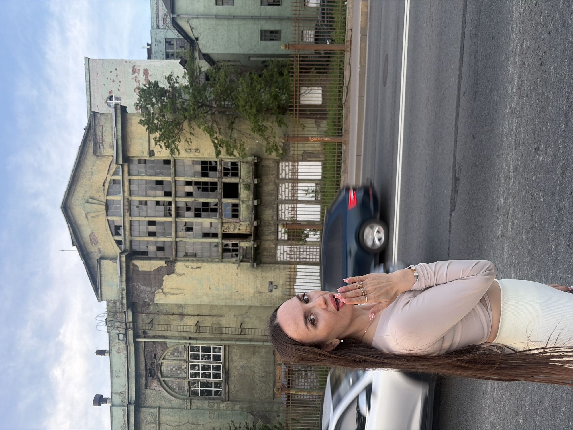 The image captures an urban street scene in Saint Petersburg, Russia, featuring a woman in the foreground against the backdrop of the dilapidated Krasny Treugolnik (Red Triangle) factory complex, specifically at the Obvodny Canal Embankment, 134-138.
In the immediate foreground, slightly right of center, a woman with long, straight dark brown hair, fair skin, and full lips with pink lipstick stands, looking directly at the viewer. She is dressed in a light beige long-sleeved cropped top and a white ribbed high-waisted skirt. Her left hand, adorned with multiple rings and a bracelet, is raised to her mouth, with her fingers partially covering it and her lips slightly ajar. Her fingernails are light pink with small embellishments.
Behind the woman, the main subject of the background is a large, multi-story industrial building from the Krasny Treugolnik complex. The building's facade is heavily weathered, exhibiting peeling pale green and yellow paint and crumbling plaster that reveals underlying brickwork. It features a prominent central section with a large, multi-paned window structure, most of which have broken glass. To the left, an arched window with an intact grid of smaller panes is visible. A metallic ladder ascends the exterior wall on the left side of the building's roofline. Further to the right, a second, less dilapidated green building, also part of the complex, is visible.
A rusty metal picket fence spans horizontally across the midground, separating the factory grounds from the street. Behind the fence, a small, leafy green tree grows. The foreground features an asphalt road with a double white line. Two vehicles are in motion on the road: a silver sedan is partially visible on the left, obscured by the woman, and a dark blue SUV is seen on the right, both blurred to indicate movement. The scene is illuminated by natural daylight, under a partly cloudy blue sky.