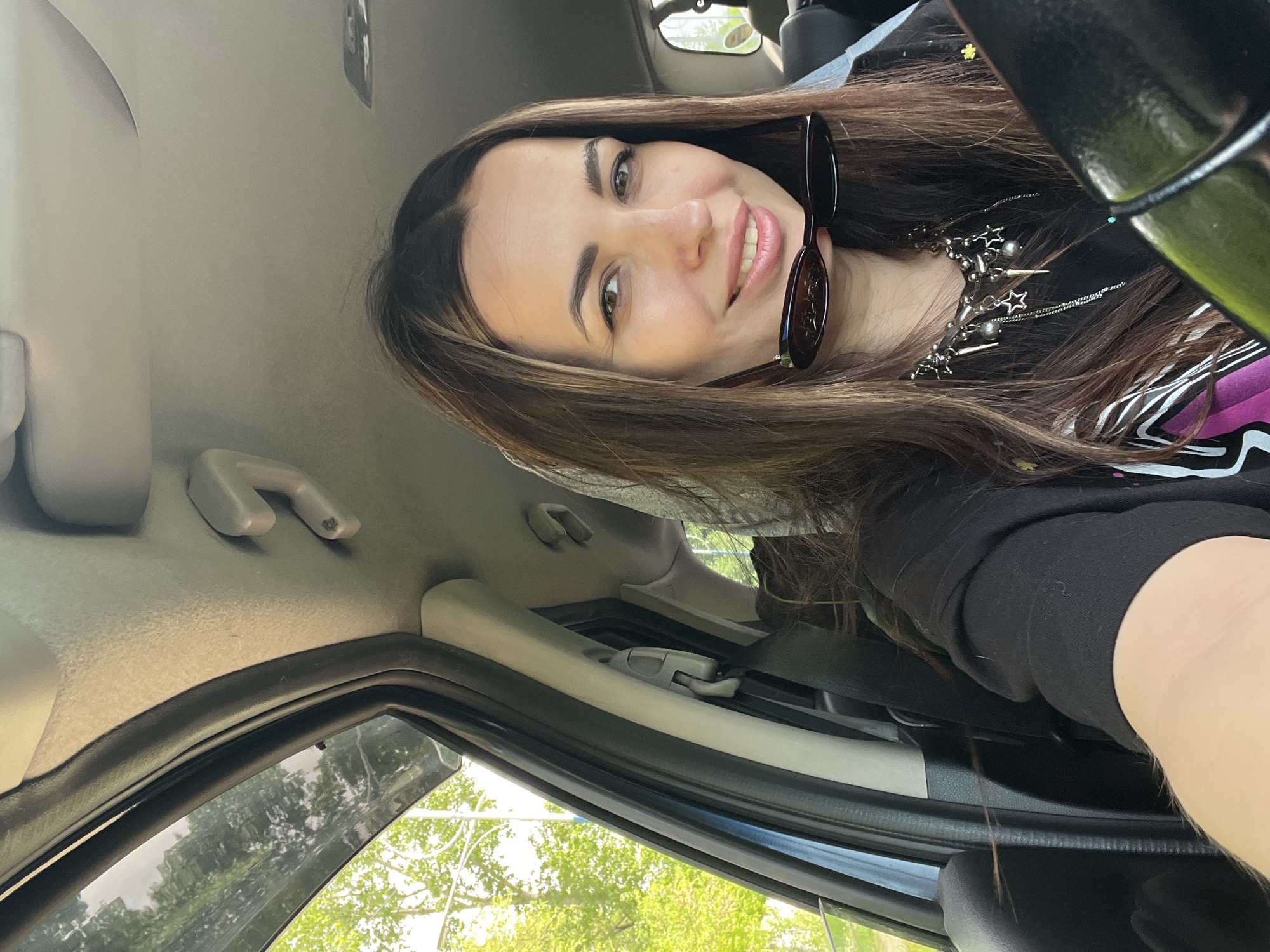 A selfie photograph shows a smiling woman inside a vehicle, likely a car, with the steering wheel visible in the top right. She is wearing a dark top, sunglasses, and a decorative necklace with star elements. The interior of the car is visible, including the dashboard, window, and ceiling. Outside the window, trees and foliage are visible, suggesting a daytime setting.