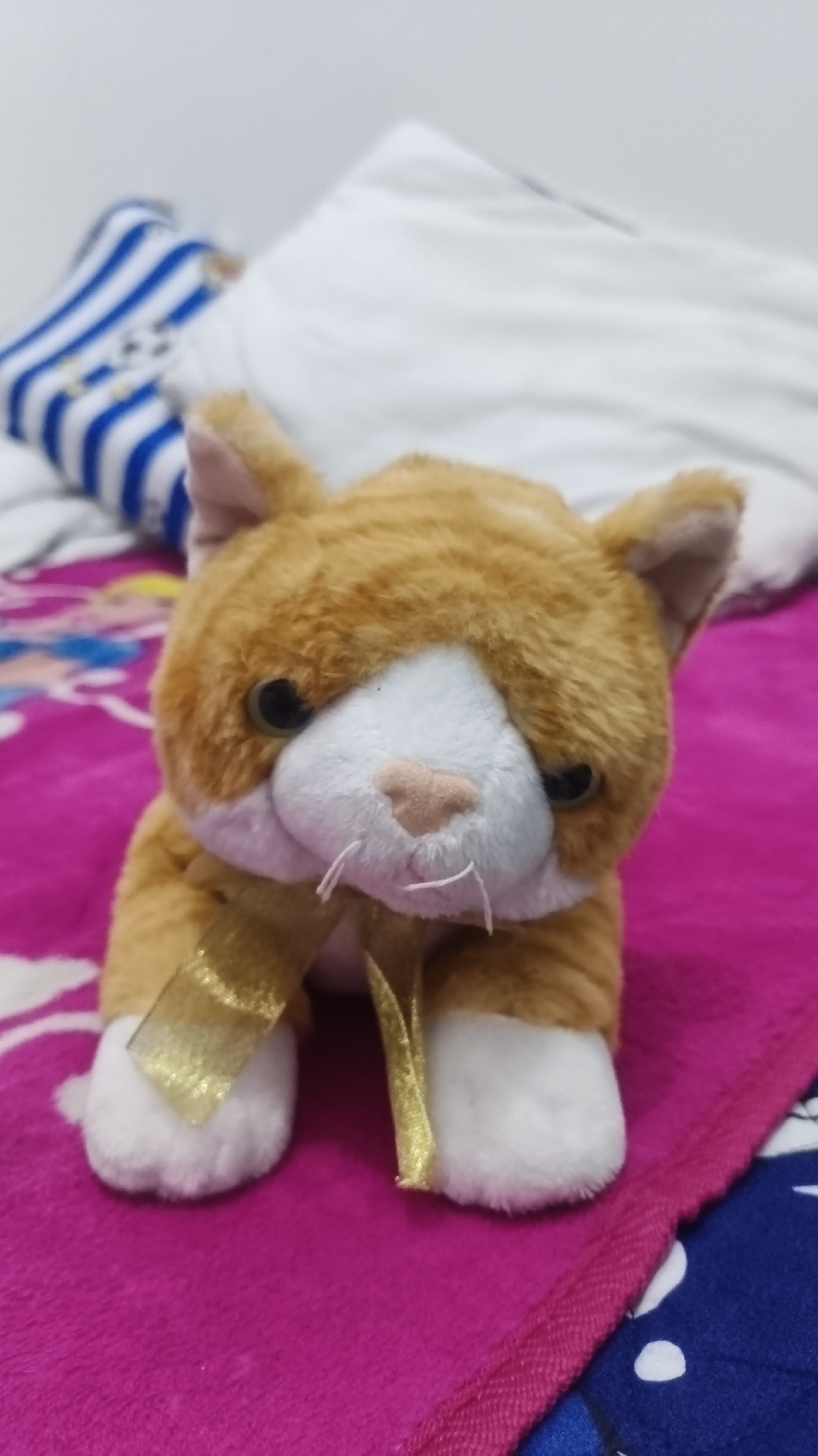 A stuffed toy cat, primarily orange and white, sits on a pink blanket. A gold ribbon is tied around its neck. In the background, pillows and a striped blanket are visible. The scene appears to be indoors, likely in a bedroom. There is no information to suggest this location is in Betung, Indonesia.
